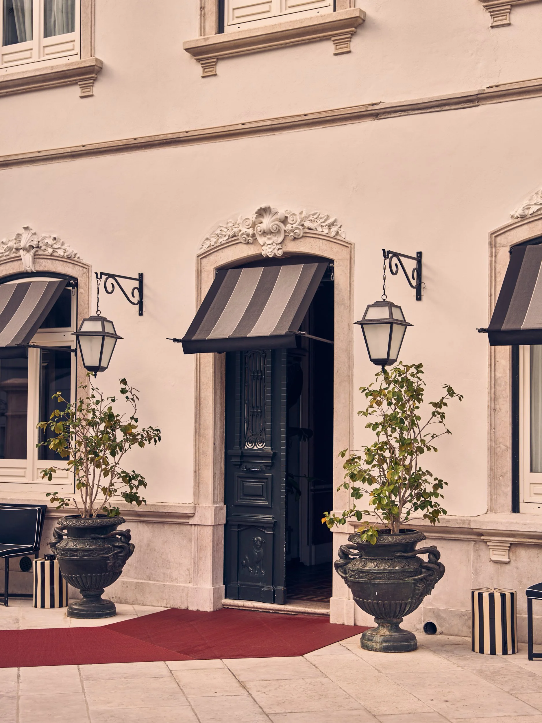 The entrance of a building with a black door, decorative stone frame, striped awnings, and two large potted trees on either side, with a red carpet on the tiled porch.