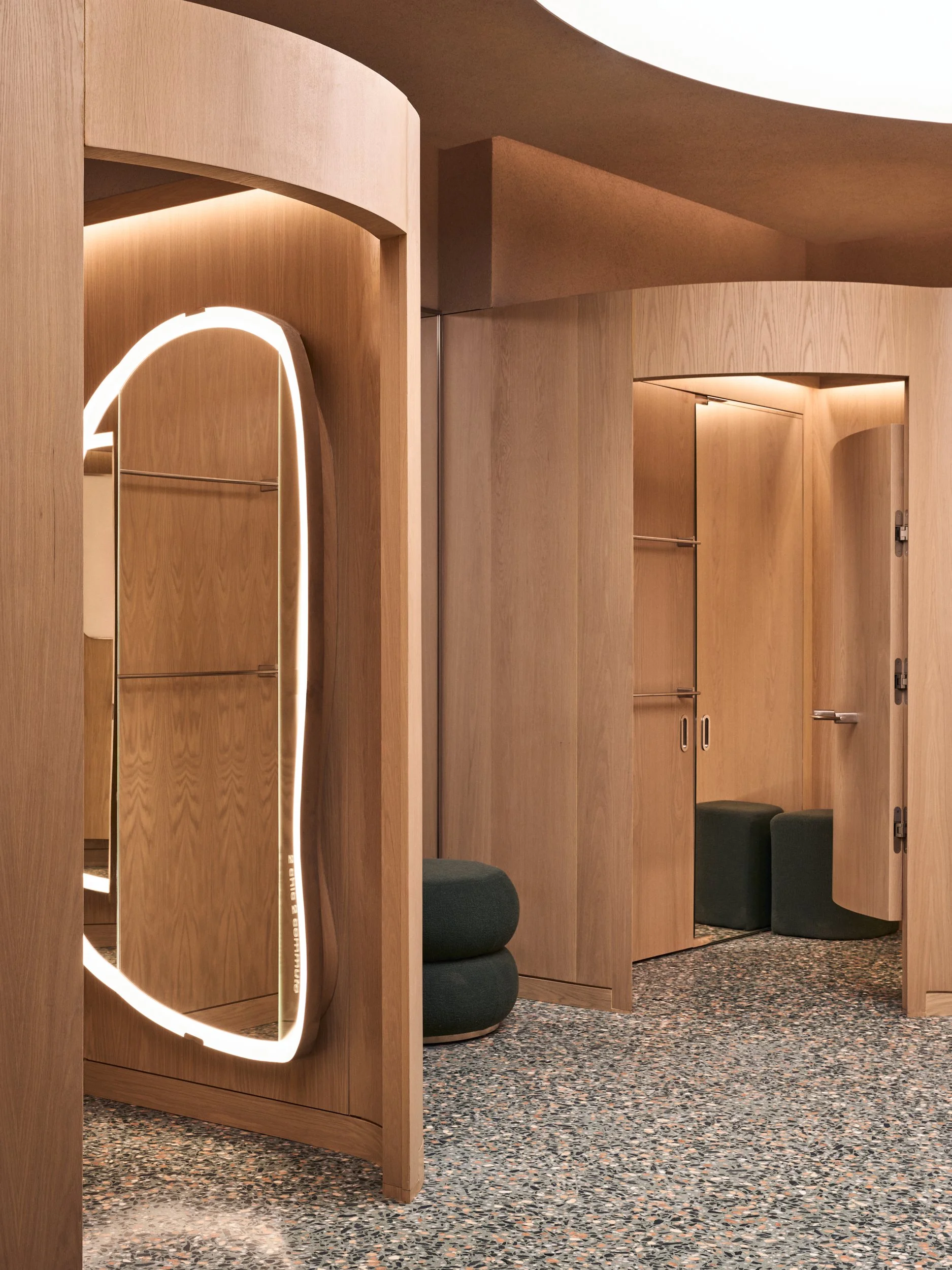 Modern retail dressing area with curved wooden walls, seating, and illuminated mirror or light fixtures, on a speckled floor.