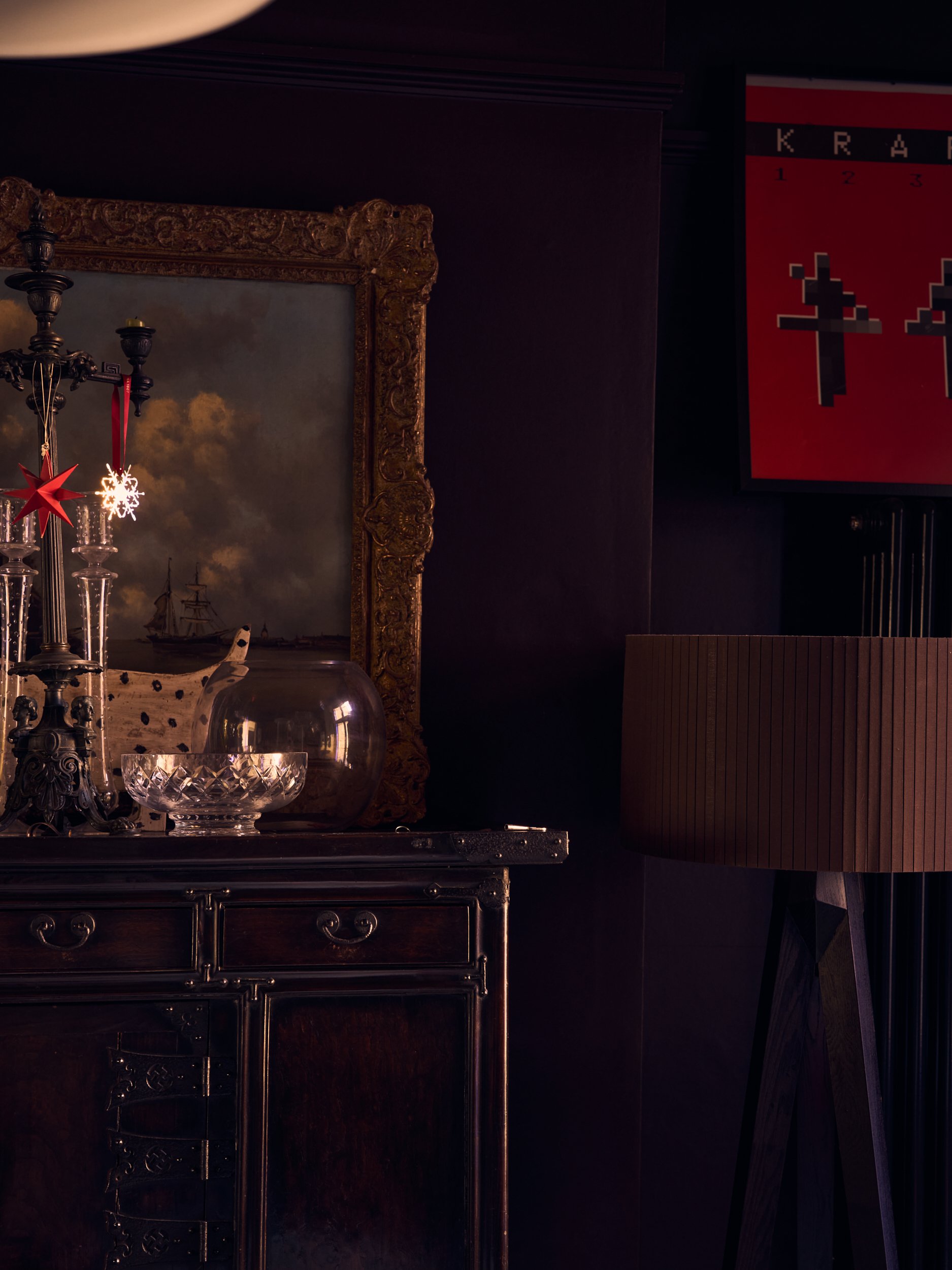 A close-up of an ornate gold-framed painting, part of a chandelier decoration with Christmas ornaments, a glass bowl, and a dark sideboard against a dark wall with a red and black poster featuring pixelated crosses and numbers.