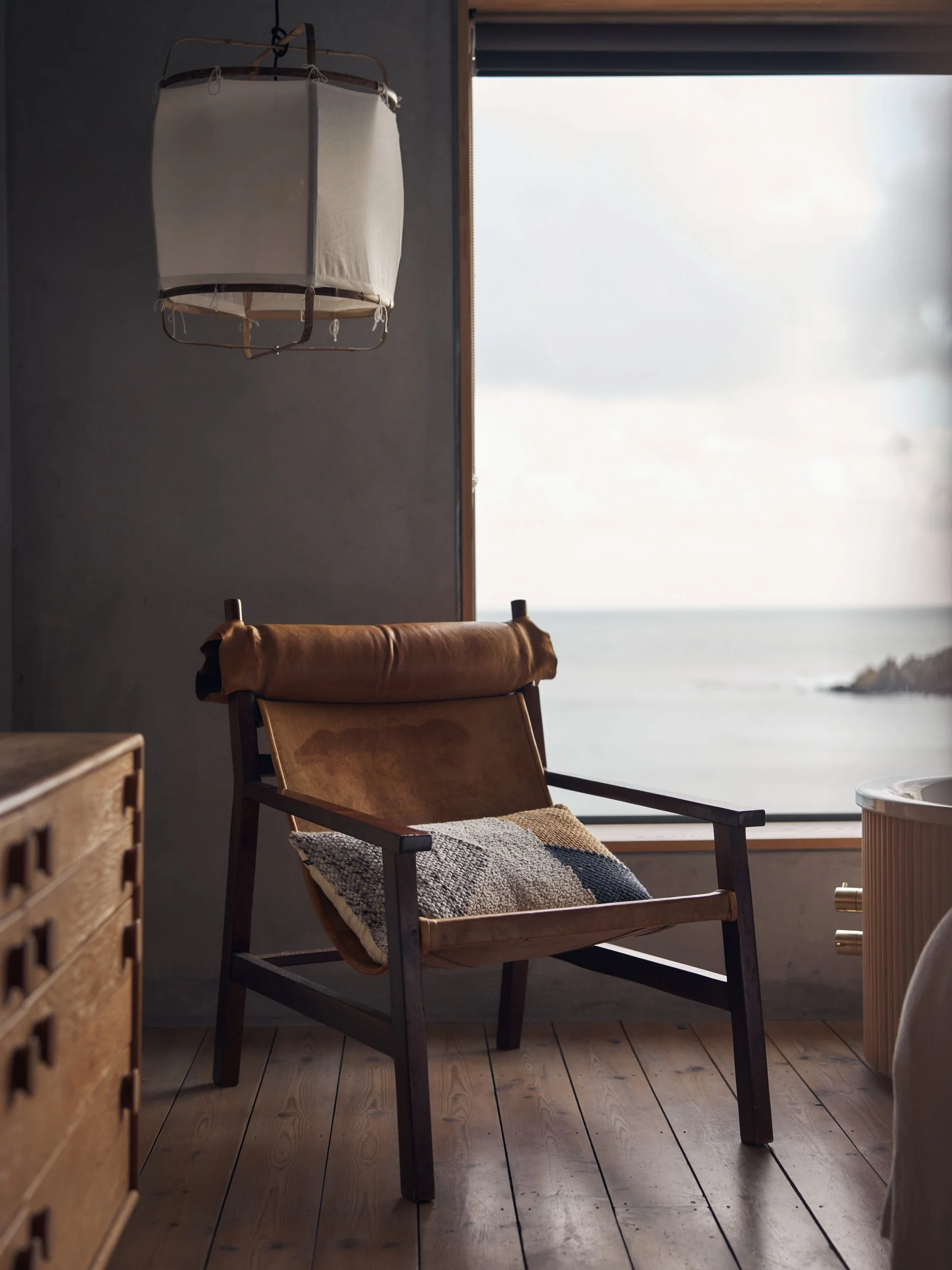 A wooden chair with a patterned pillow, positioned near a large window overlooking the ocean, with a hanging lampshade above and a small round table nearby.