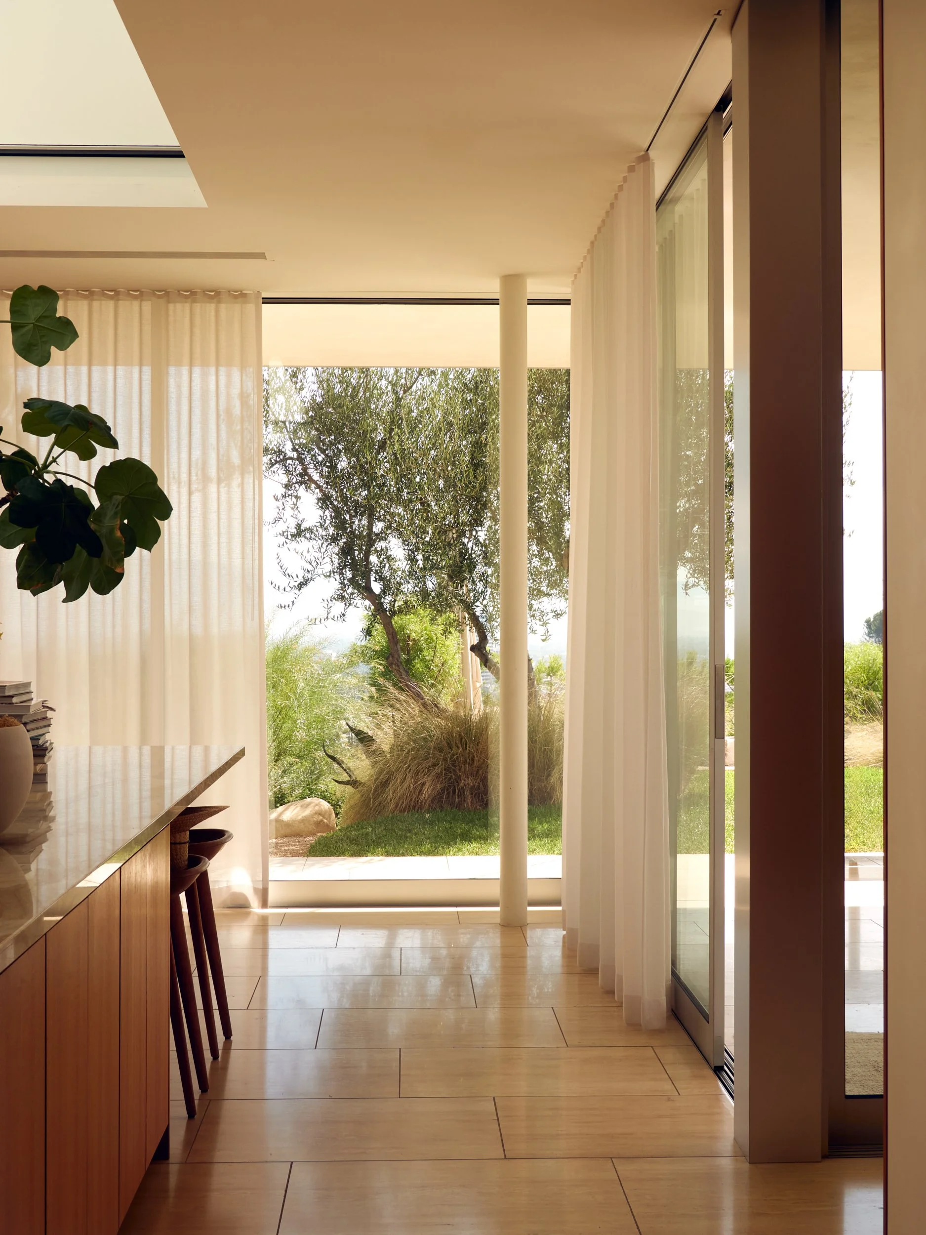 A modern interior room with large glass sliding doors opening to a green outdoor landscape with trees and bushes, white curtains, wooden furniture, and light-colored tiled flooring.