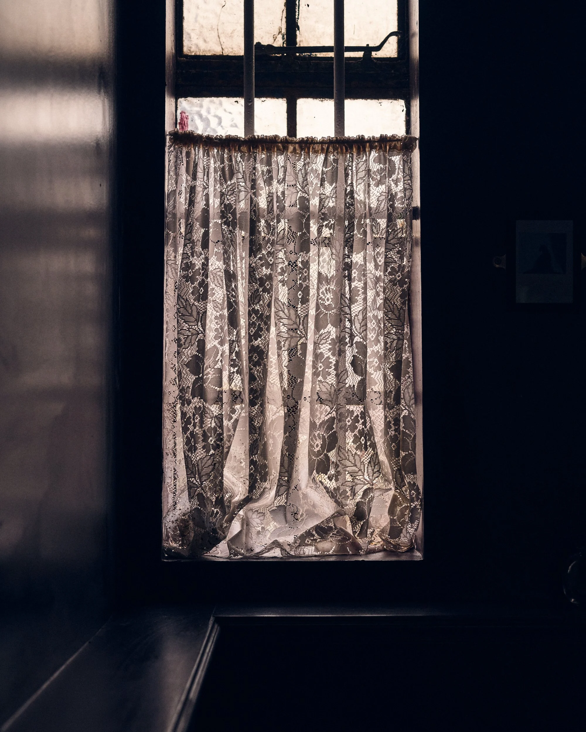 A window with lace curtains seen through a dark frame, with sunlight coming through the window.