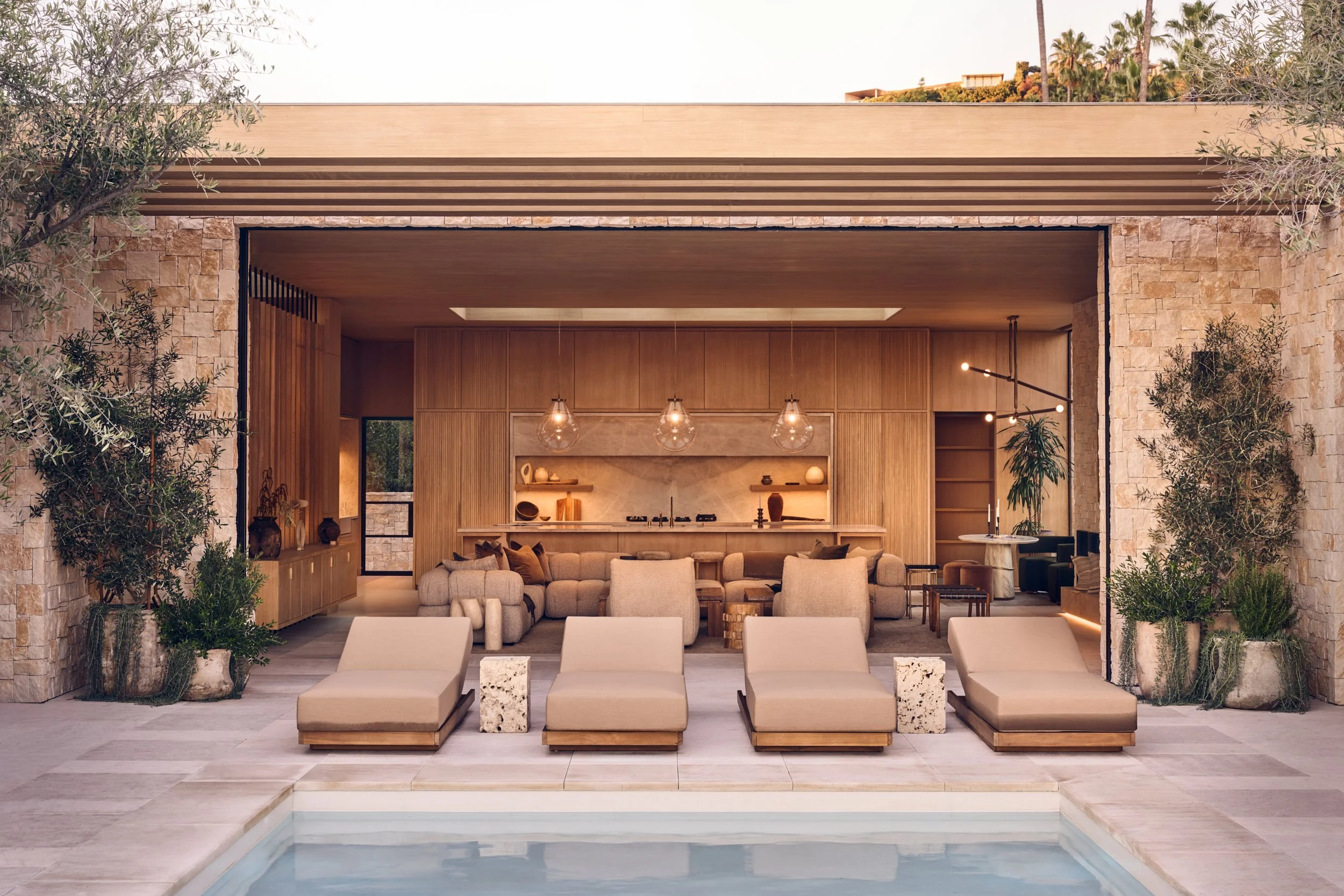 Exterior view of a modern house with earthy stone walls, wooden accents, and a spacious seating area with beige cushioned chairs and a small pool in the foreground, surrounded by trees and palm trees.