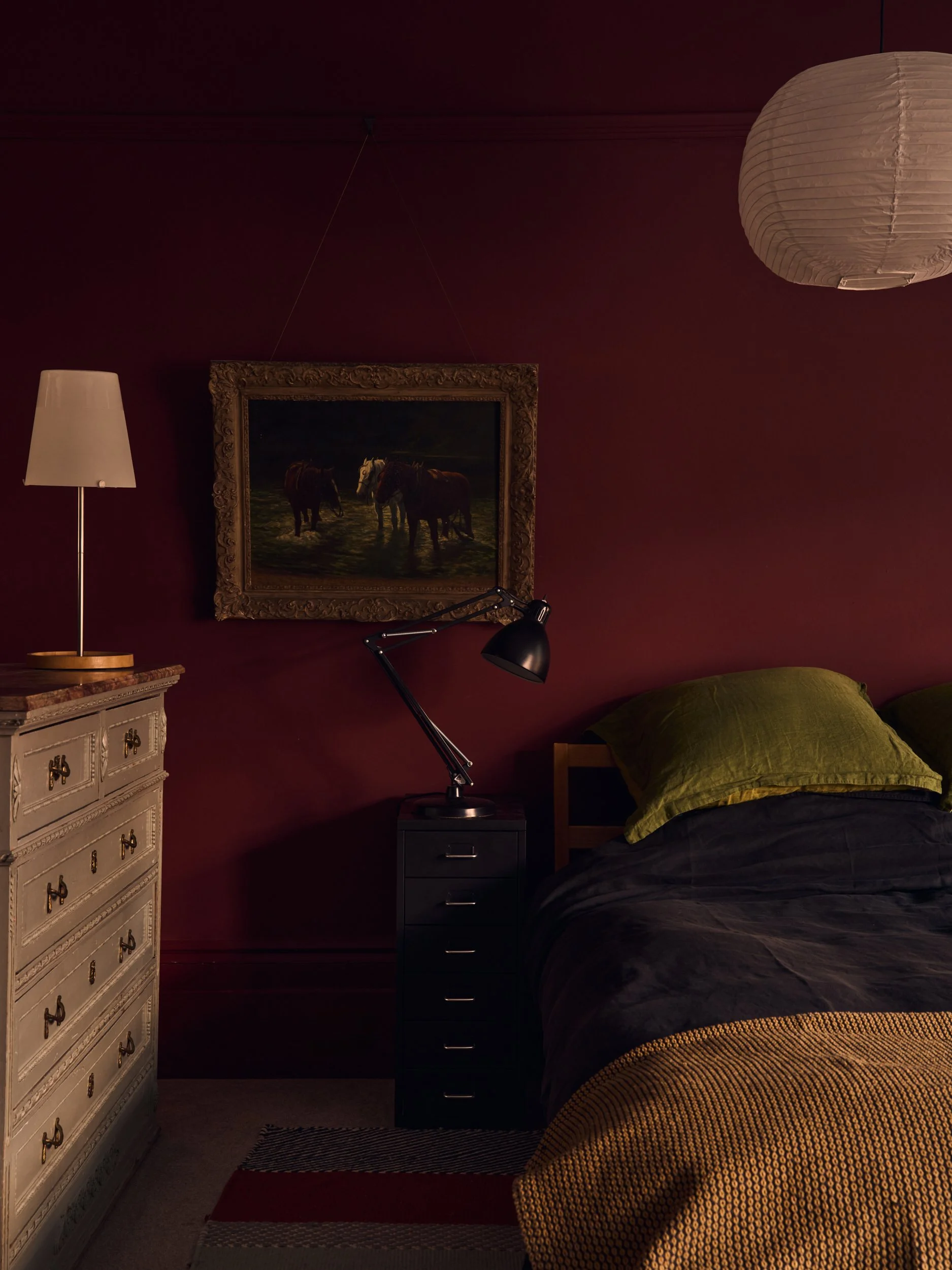 A bedroom with dark red walls, a white dresser, a black bedside table with a lamp, a framed painting of horses on the wall, a bed with green pillows and a black and yellow striped blanket, a hanging paper lantern, and a desk lamp on the bedside table
