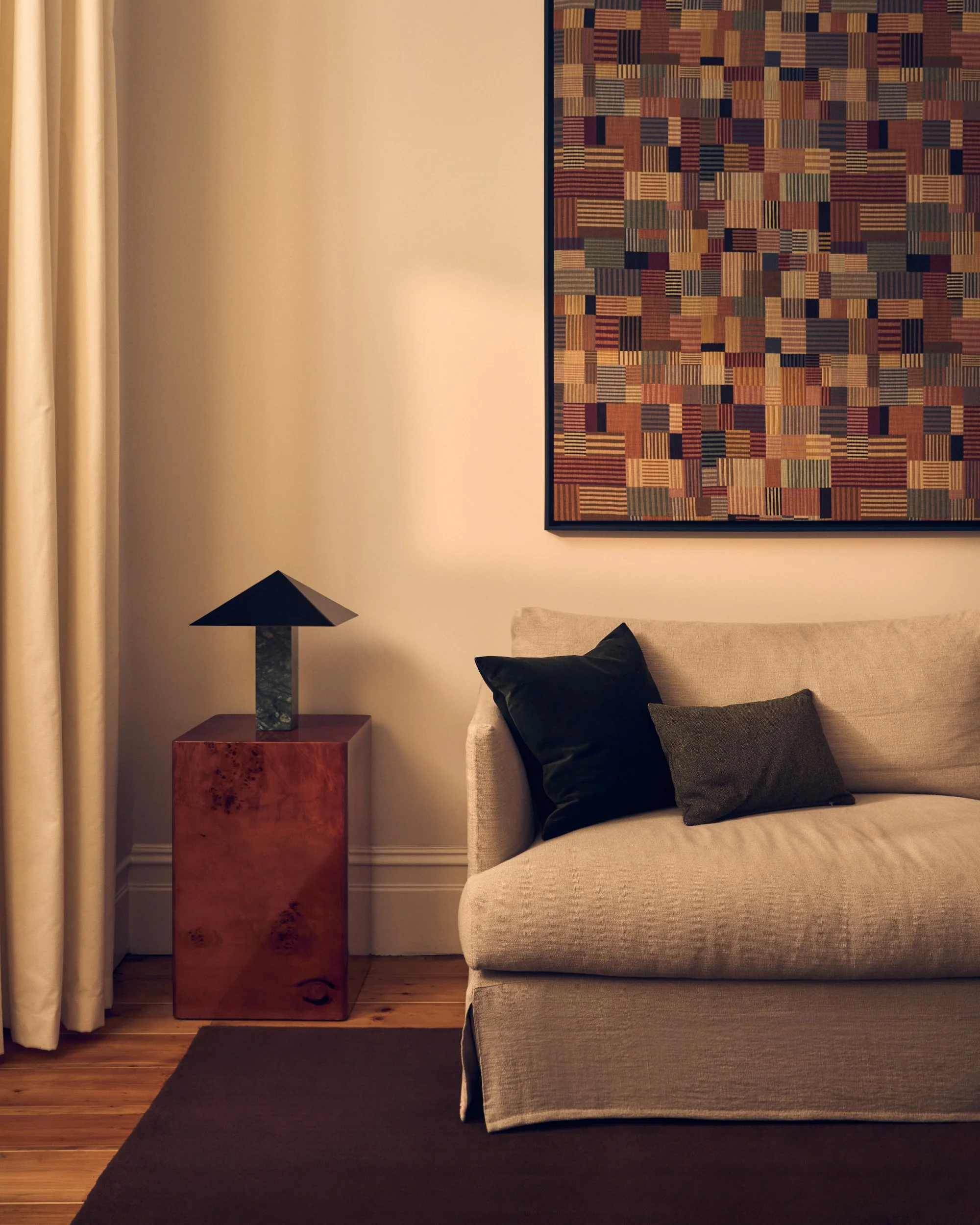 A living room corner with beige sofa, black and green throw pillows, wooden side table holding a contemporary table lamp, abstract geometric artwork on the wall, cream curtains, wooden floor, dark area rug.