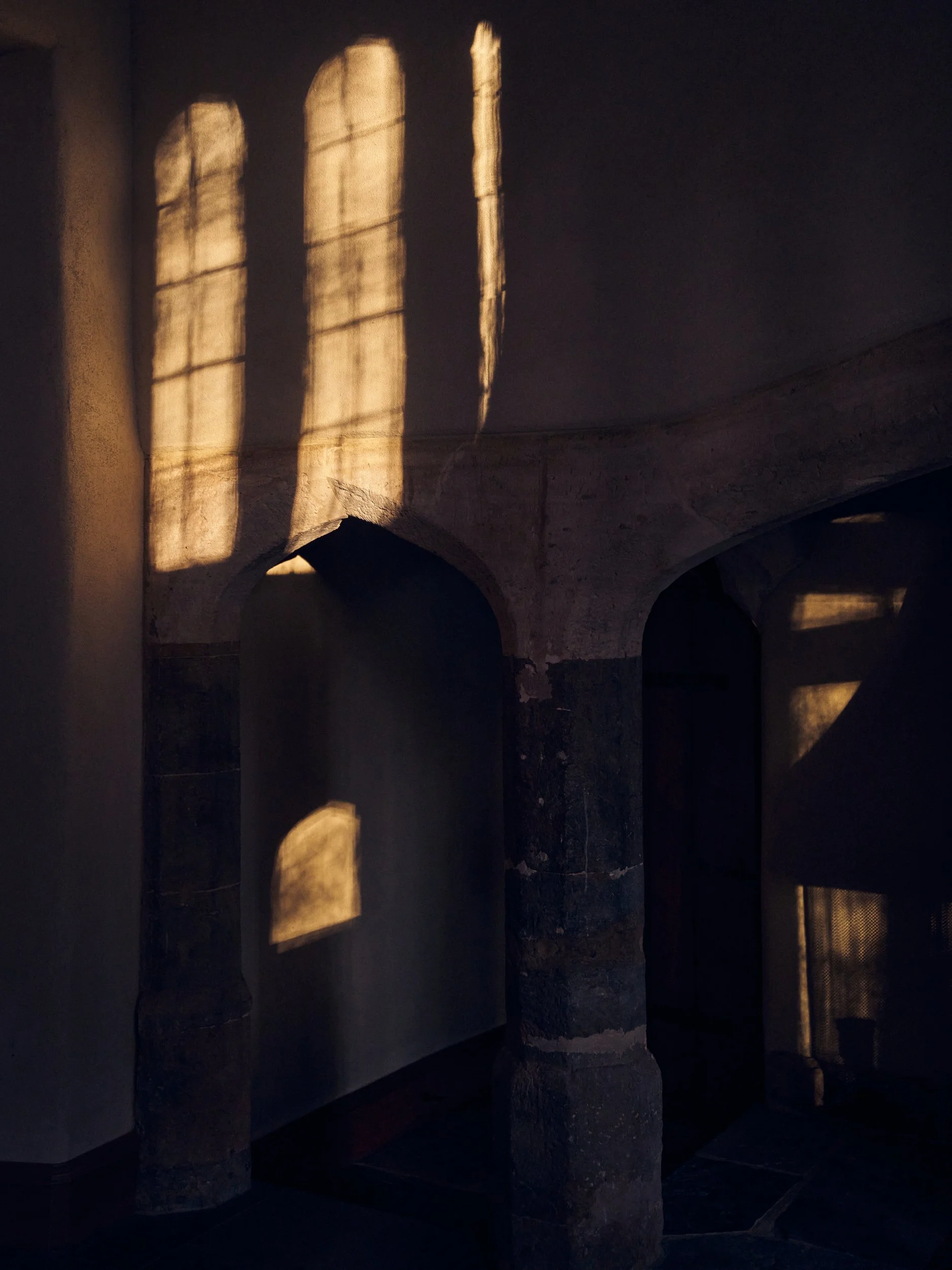 Sunlight coming through arched windows and casting shadows on a stone wall inside a building.