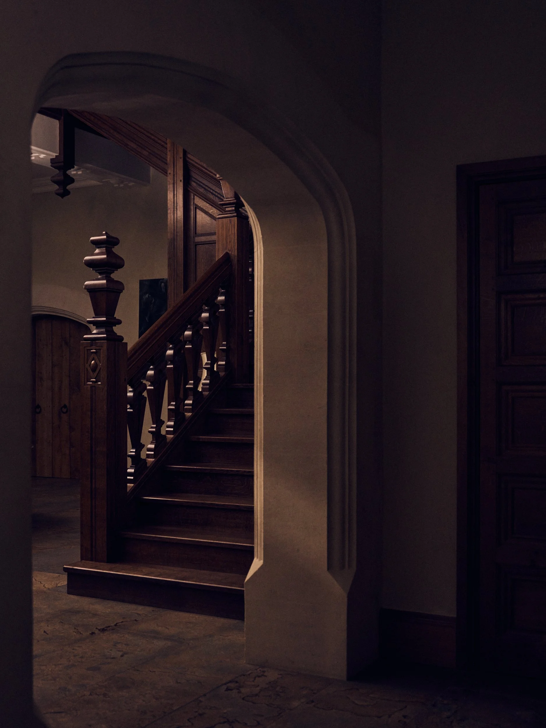 A dimly lit staircase with wooden railings and a decorative newel post, seen through an archway in a dark interior.