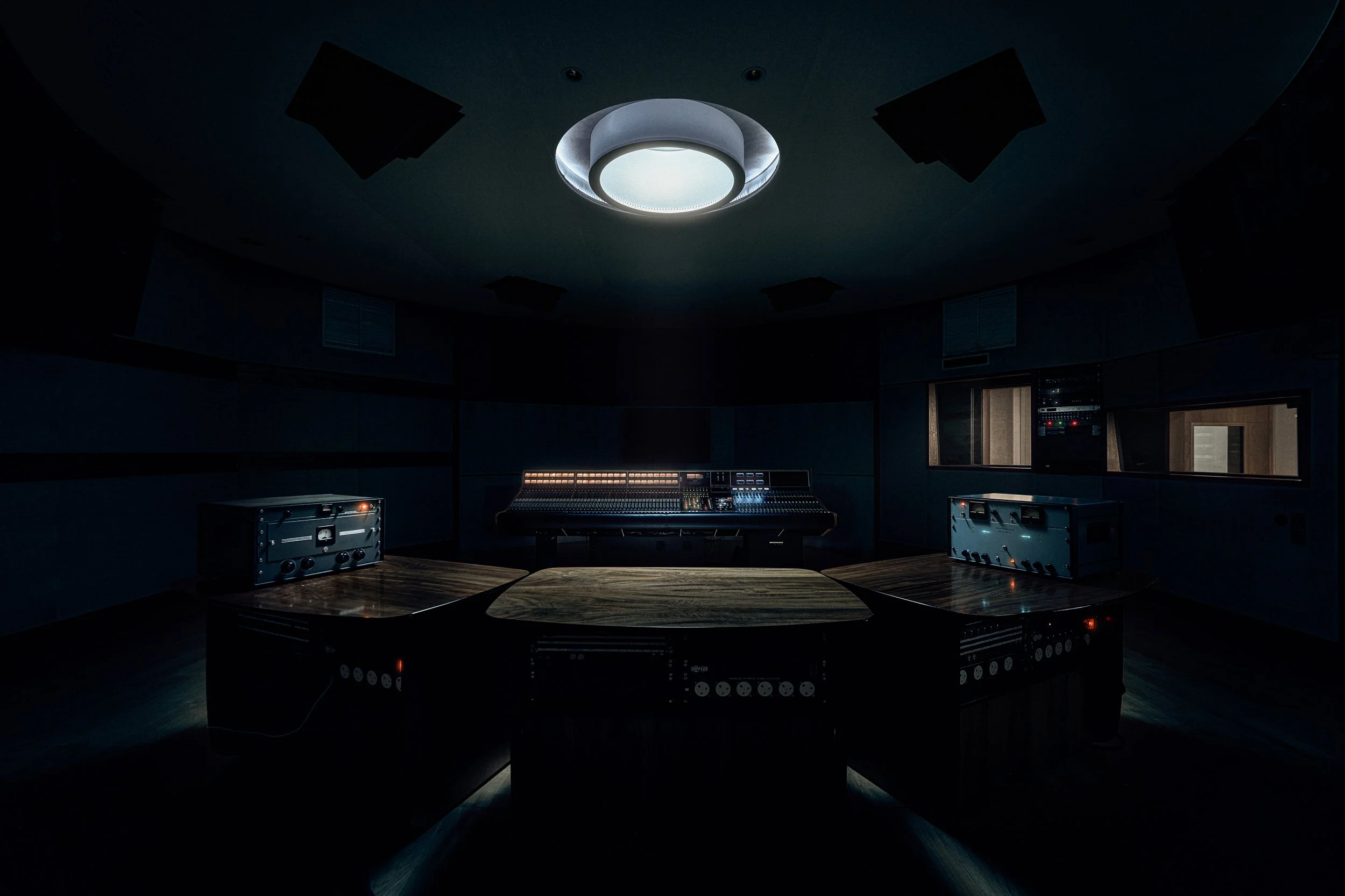 Dark recording studio with a wooden table in the foreground, a mixing console in the center, and audio equipment on the sides. The ceiling has a large round light and black soundproofing panels.
