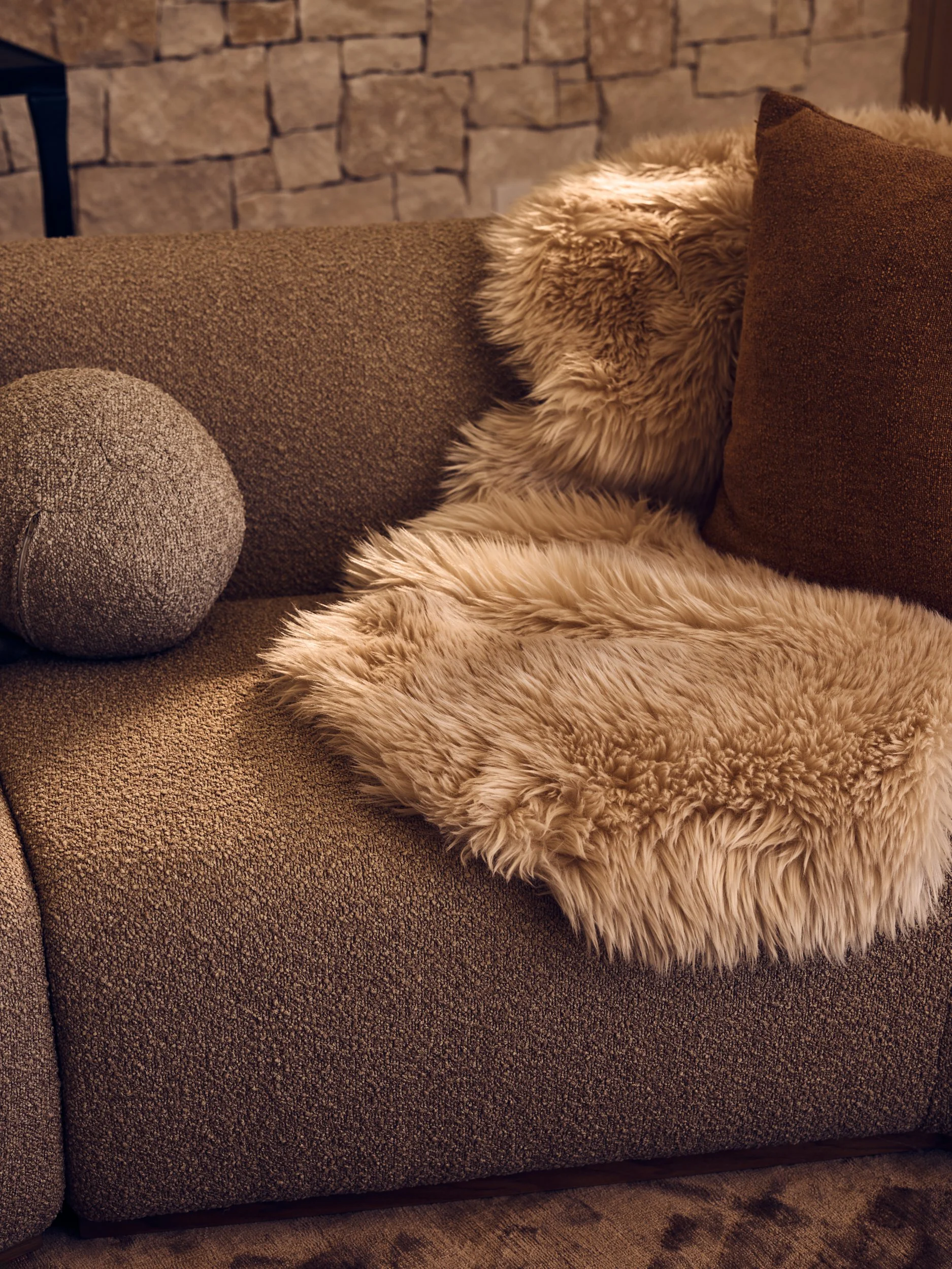 Close-up of a brown couch with a round textured gray pillow, a fluffy beige blanket, and a dark brown pillow against a brick wall.