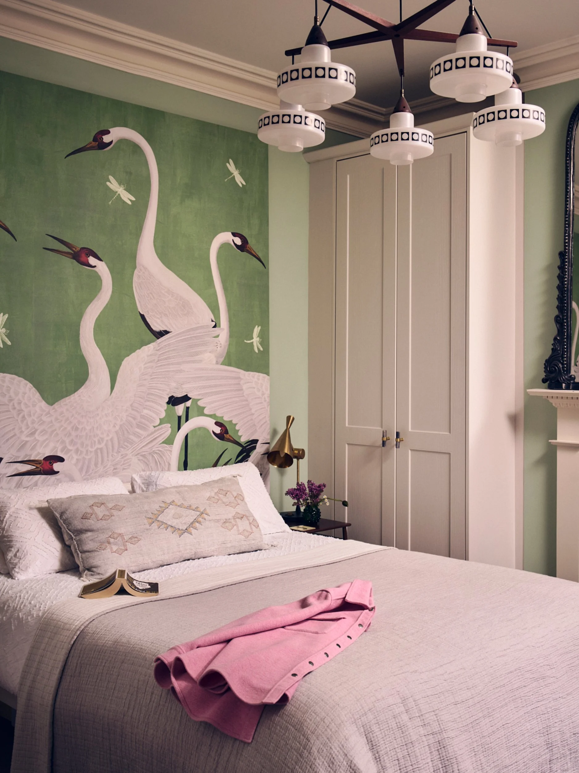 A bedroom with a bed, a pink shirt folded on the bed, a decorative pillow, a nightstand with a small bouquet of purple flowers, a brass lamp, a wardrobe, and a large wall art featuring white cranes and dragonflies on a green background. The ceiling h