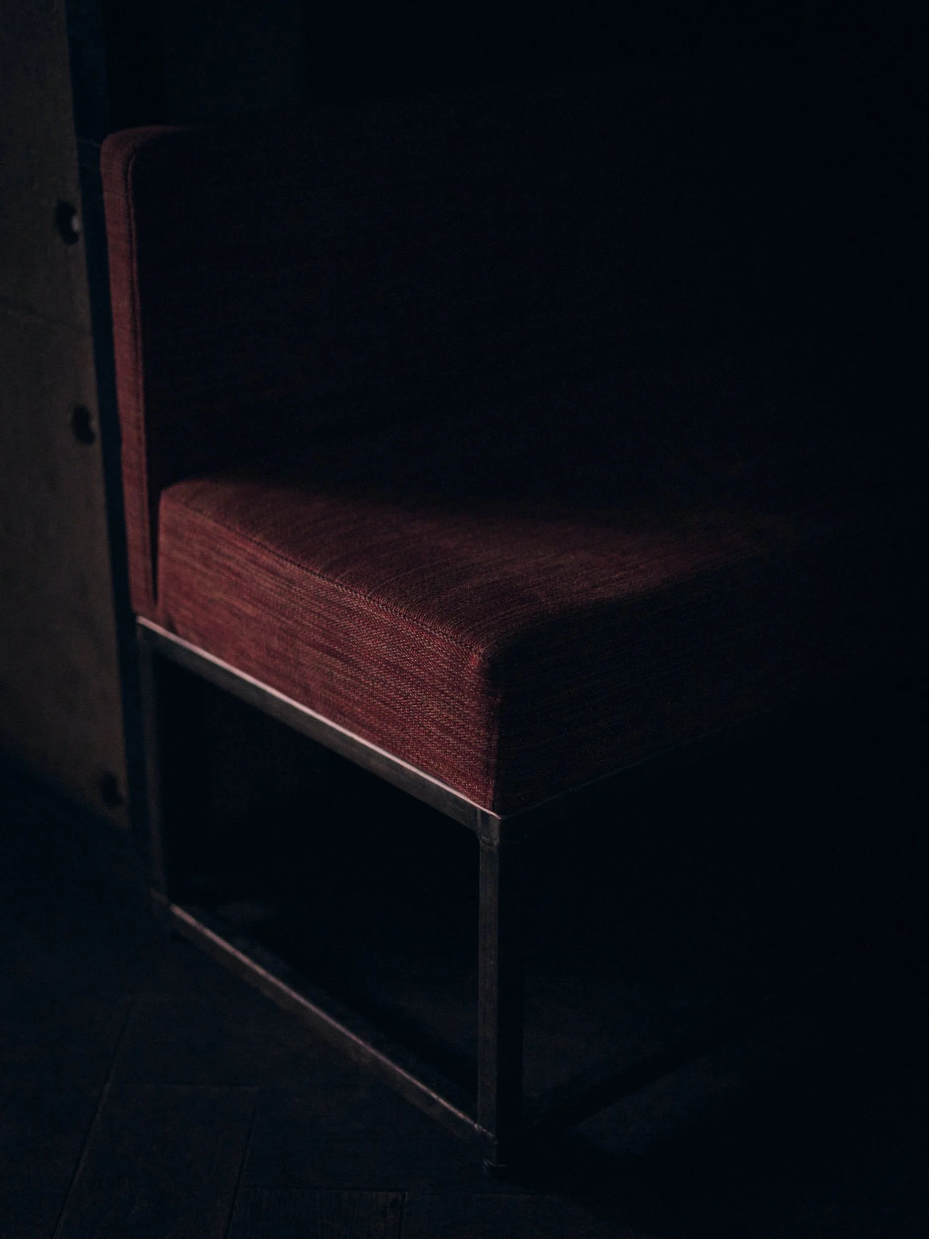 A dark room with a single red and black striped cushioned seat with metal legs. The seat is partially illuminated by minimal lighting.