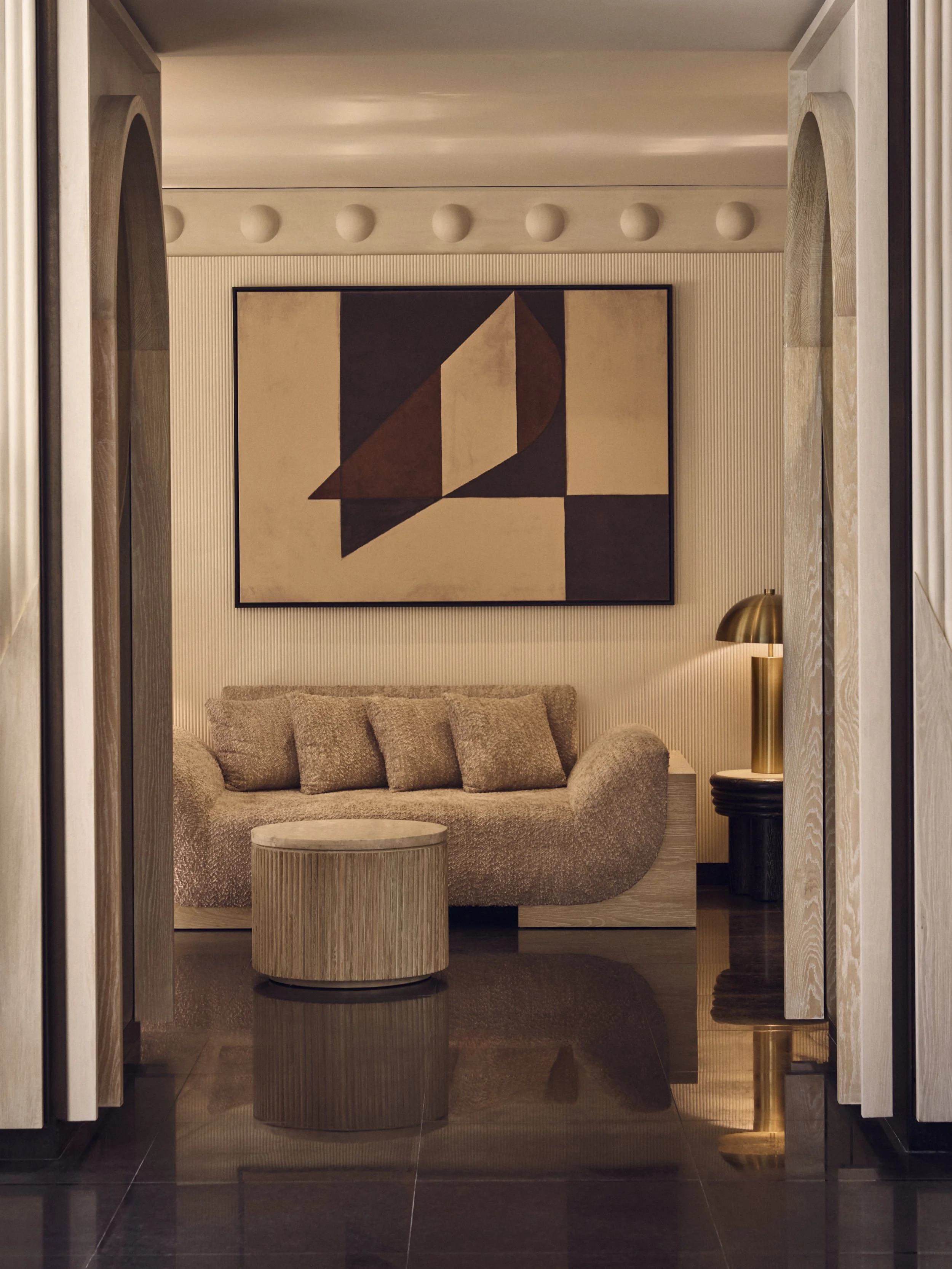 A modern living room with a textured beige sofa, a matching round wooden coffee table, a floor lamp with a gold shade, and a large abstract geometric painting on the wall.