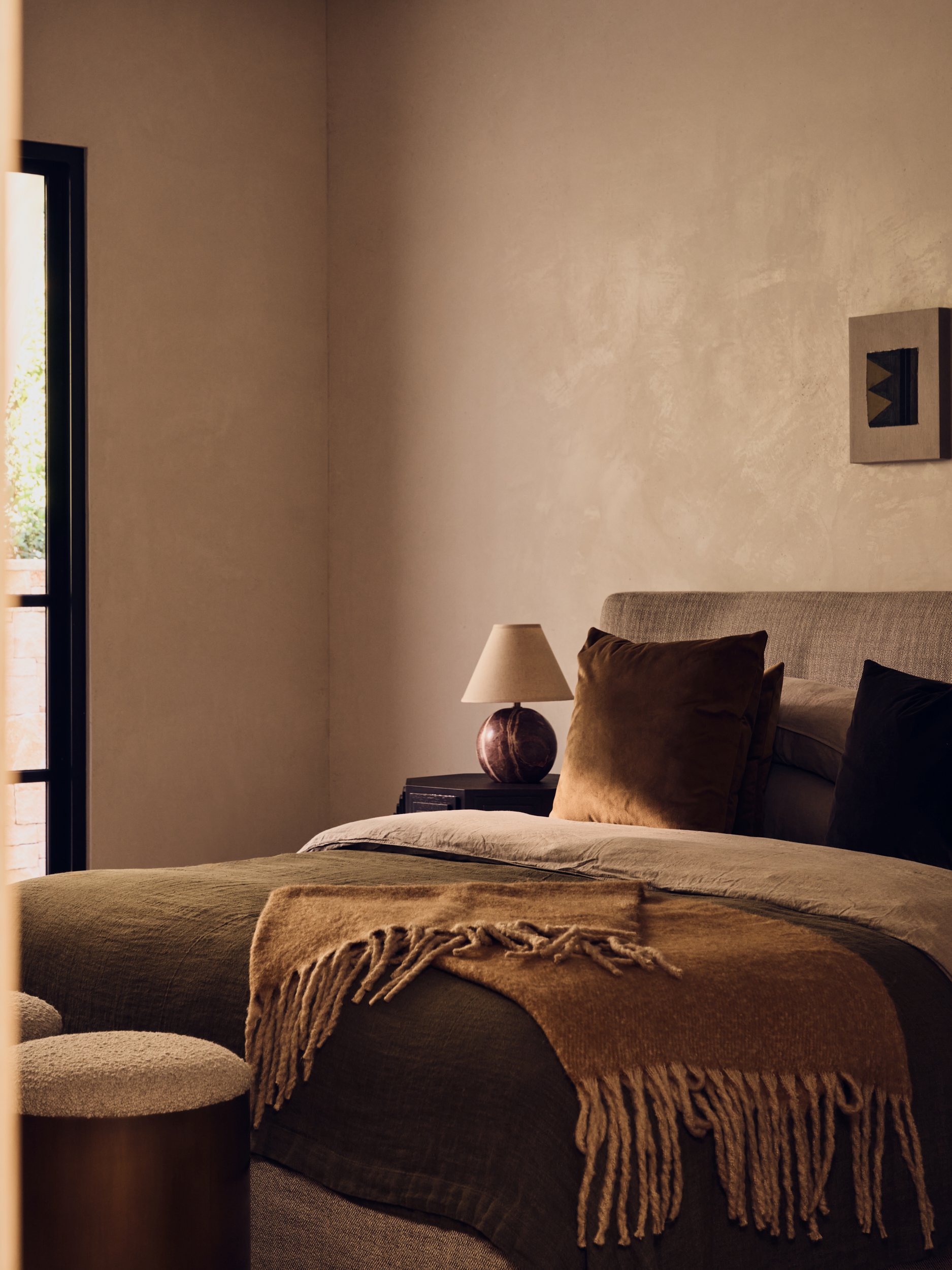 A bedroom with a bed covered in beige and brown bedding, a blanket with fringes, a side table with a lamp, and a window with sunlight streaming in.