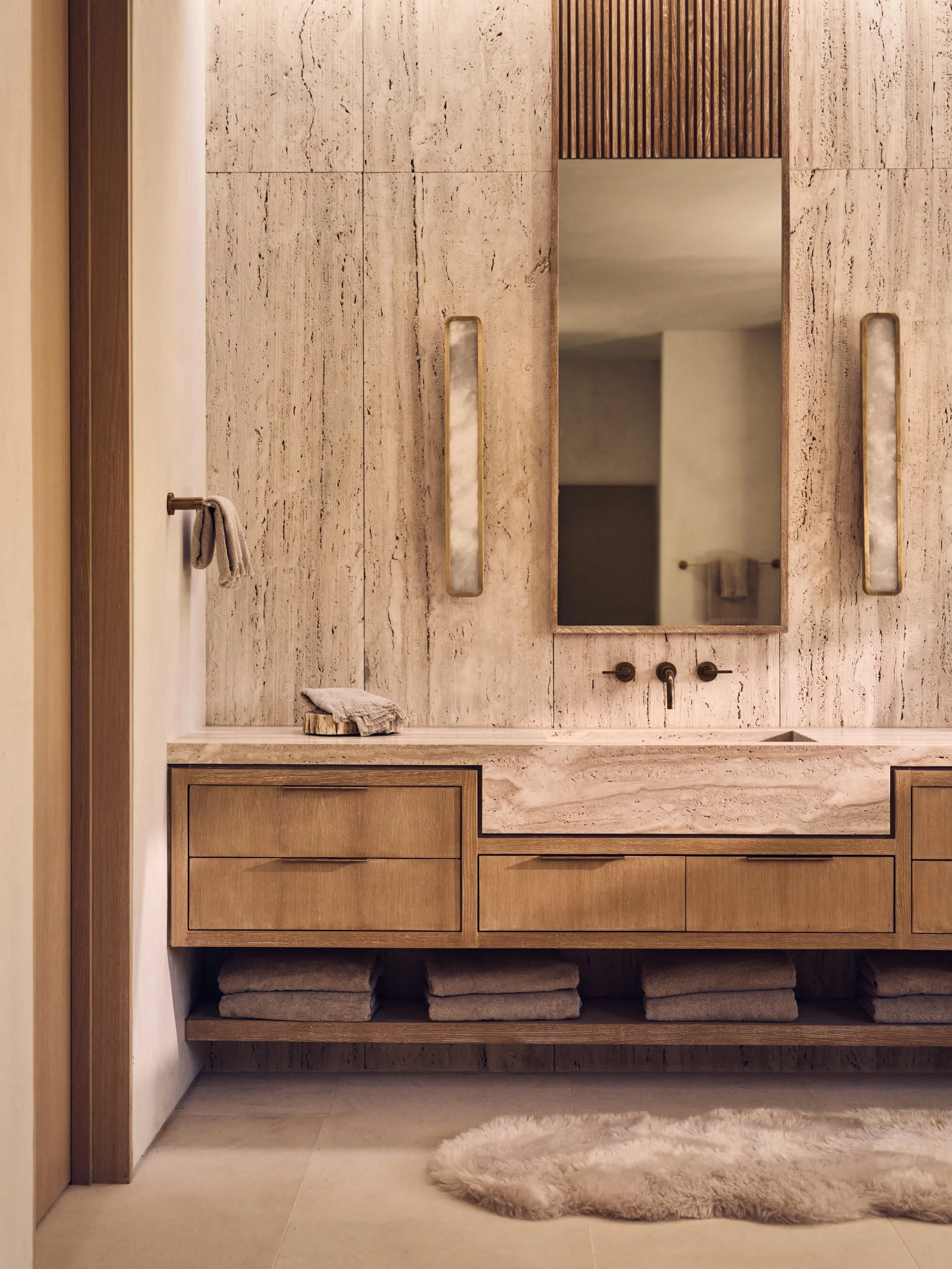 A bathroom with beige marble wall tiles, a mirror, towel rails, and a wooden cabinet with towels stored underneath. The area has neutral tones and a plush circular rug on the floor.