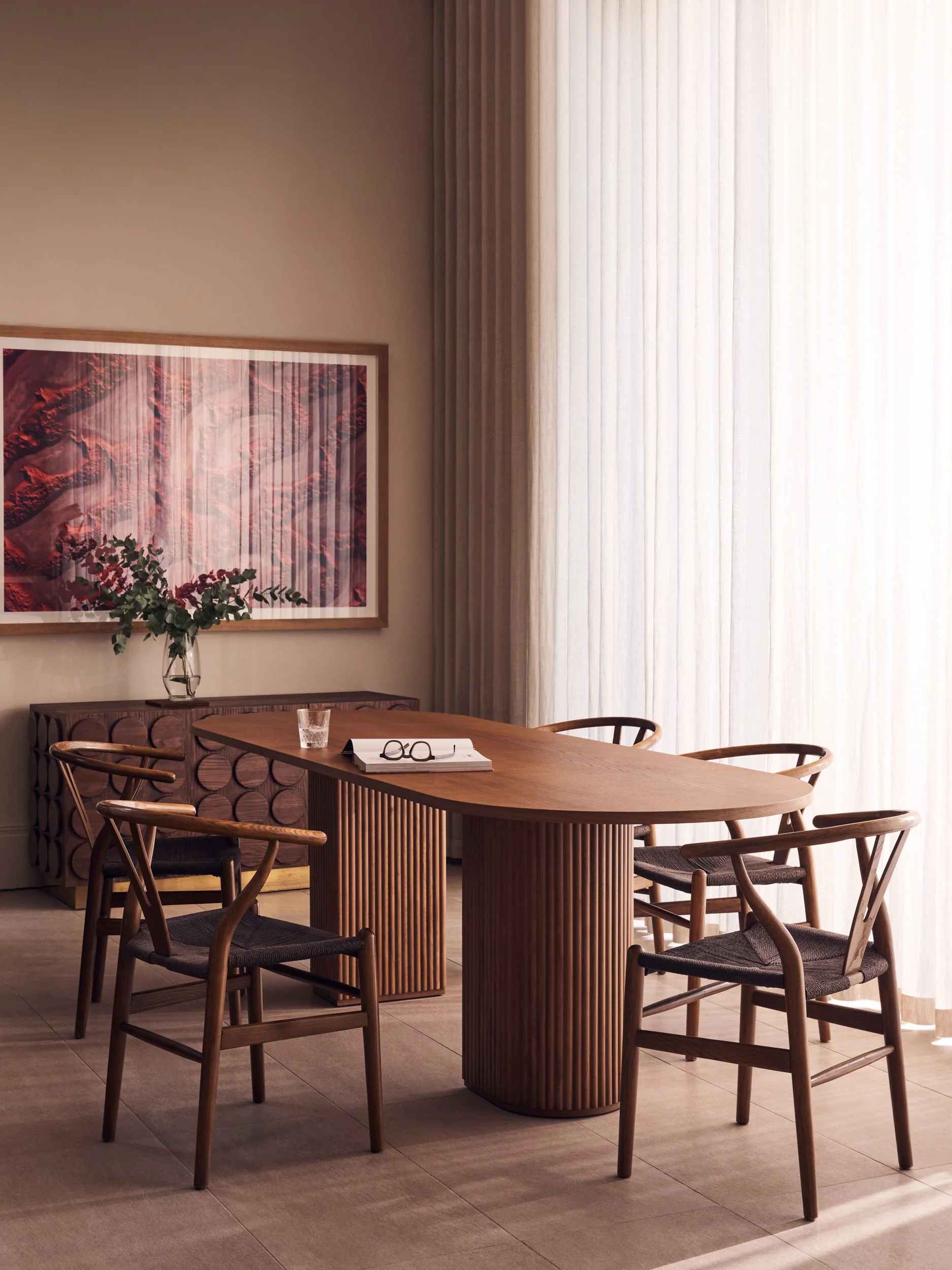 A dining room with a wooden table, six wooden chairs, a framed abstract artwork on the wall, and a tall window with sheer curtains allowing natural light, with a vase and glasses on the table.