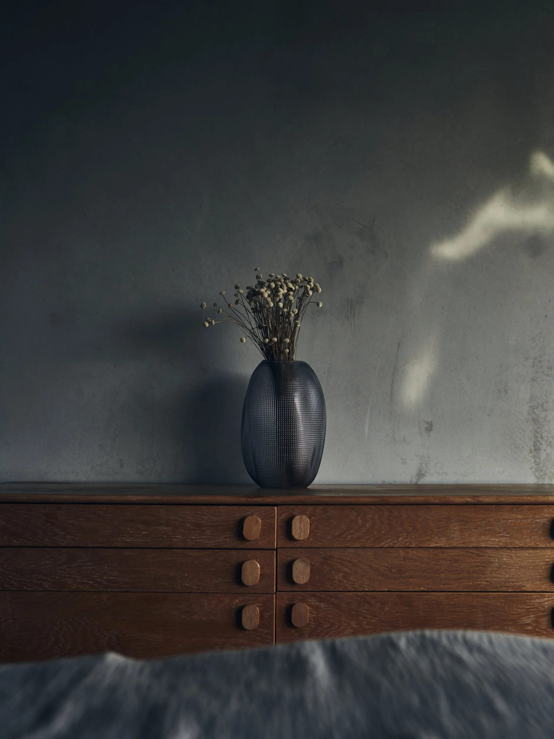 A dark-colored vase holding dried white flower stems placed on a wooden surface against a textured gray wall, with shadows and light creating a soft, muted ambiance.