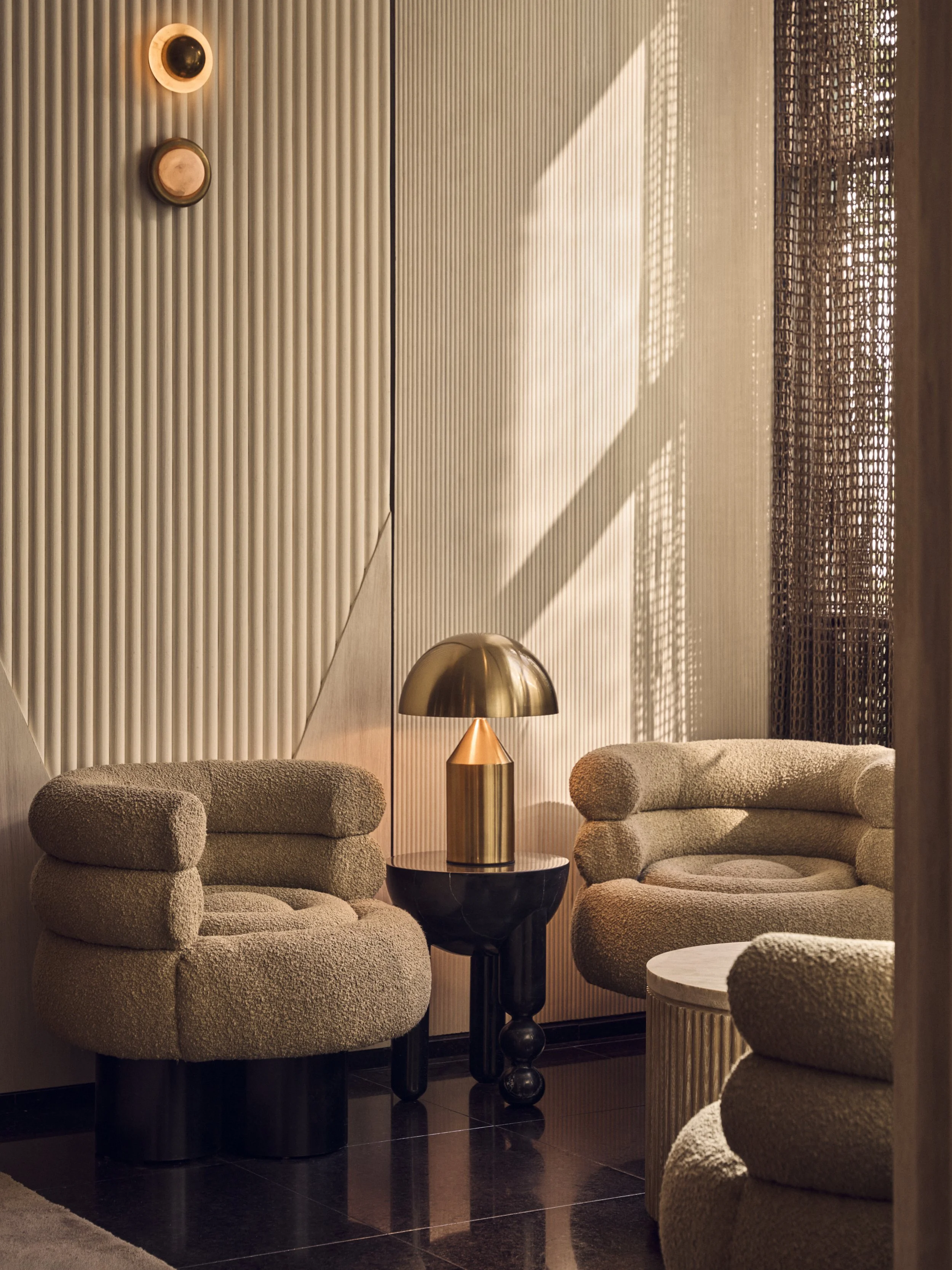 A warmly lit interior space featuring two beige plush armchairs with rounded armrests, a small round side table with a grey top, and a gold-colored table lamp with a dome-shaped shade. The background has textured vertical wall panels, a decorative wo