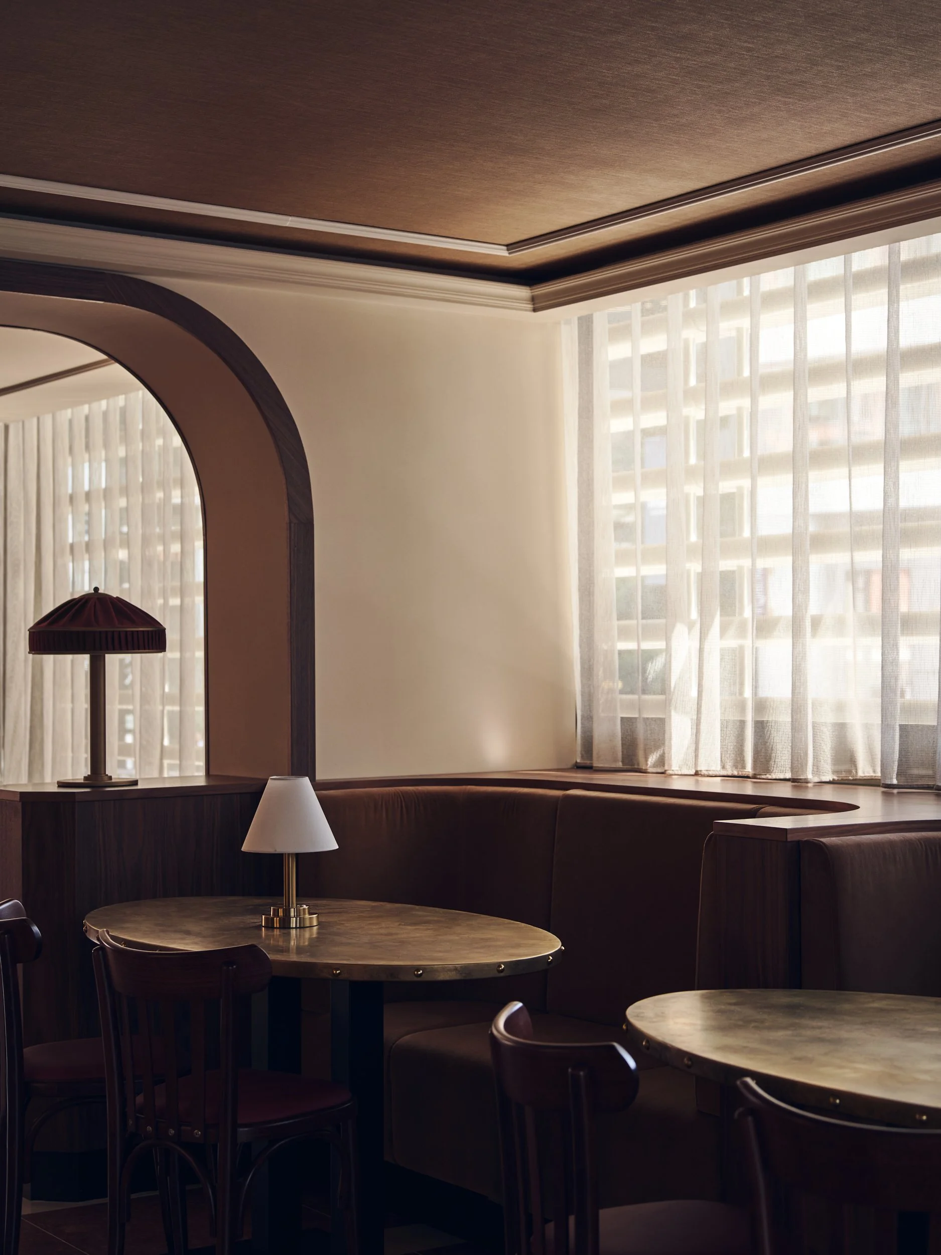 Interior of a room with round tables and chairs, large window with sheer curtains, and lamps on the tables, providing a warm and cozy atmosphere.