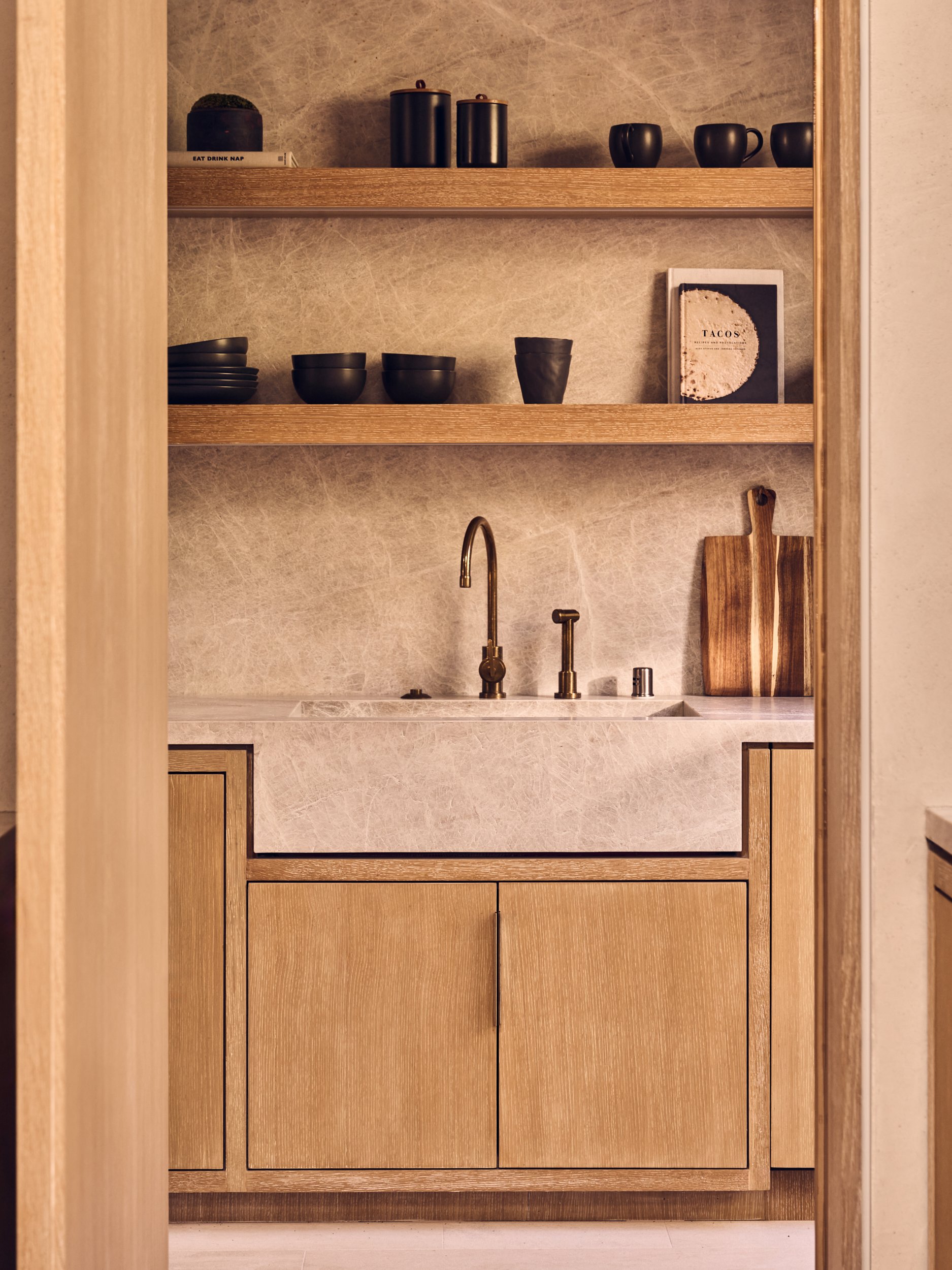 A bathroom vanity with a marble countertop, a gold-finished faucet, and wooden cabinetry. Two wooden cutting boards lean against the wall above the counter. Open wooden shelves above hold black dishes and cups, a book, and spice jars.