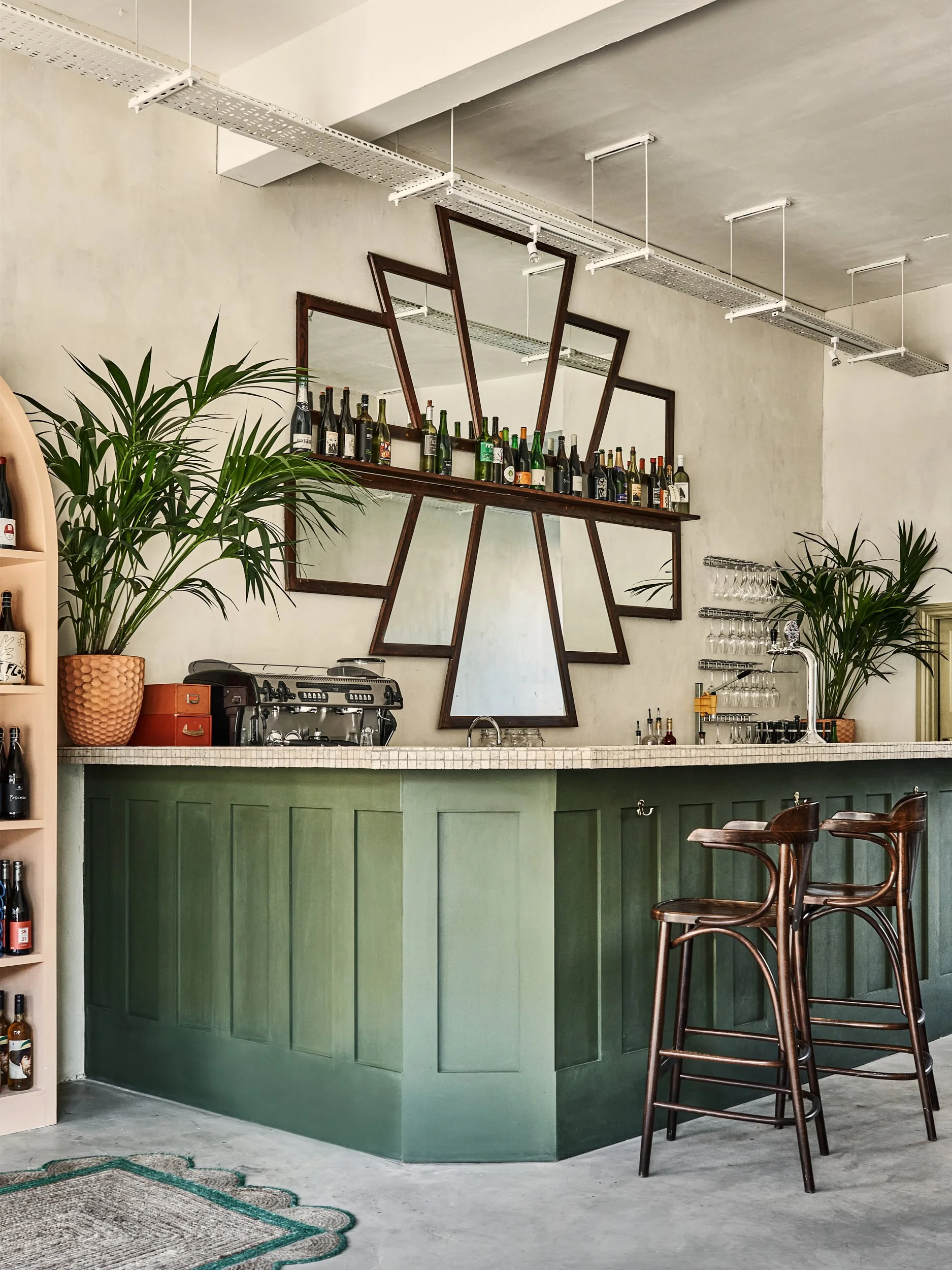A bar area with green paneling, a countertop with a coffee machine and glassware, and a large mirror with geometric wooden frames. There are two wooden bar stools with metal legs, potted plants, and a shelf with wine bottles.