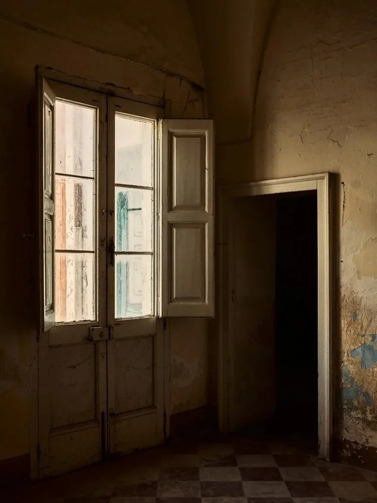 An old, abandoned room with a window and an open shutter, peeling walls, and a checkered floor.