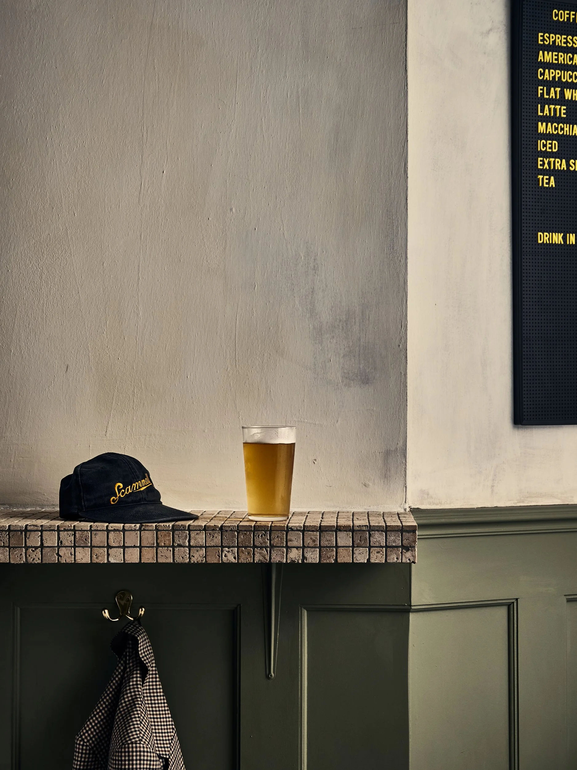 A black baseball cap with yellow embroidered text reading 'Scamall' on a wooden shelf, a glass of beer, and a black and white checkered cloth hanging from a hook on a green cabinet door. A beige and white wall in the background with a menu board part