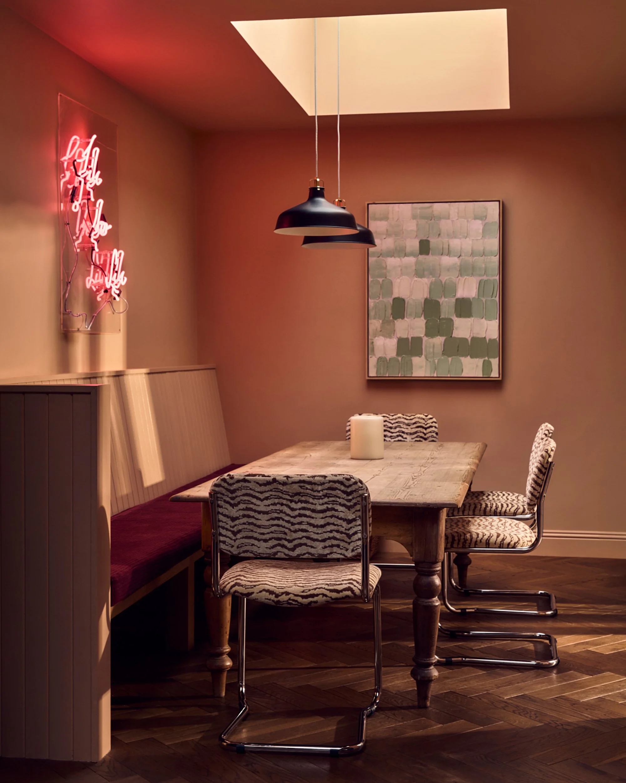 A dining area with a wooden table, mixed chairs including zebra-print, a built-in bench with red upholstery, framed wall art, black pendant lights, and a ceiling skylight.