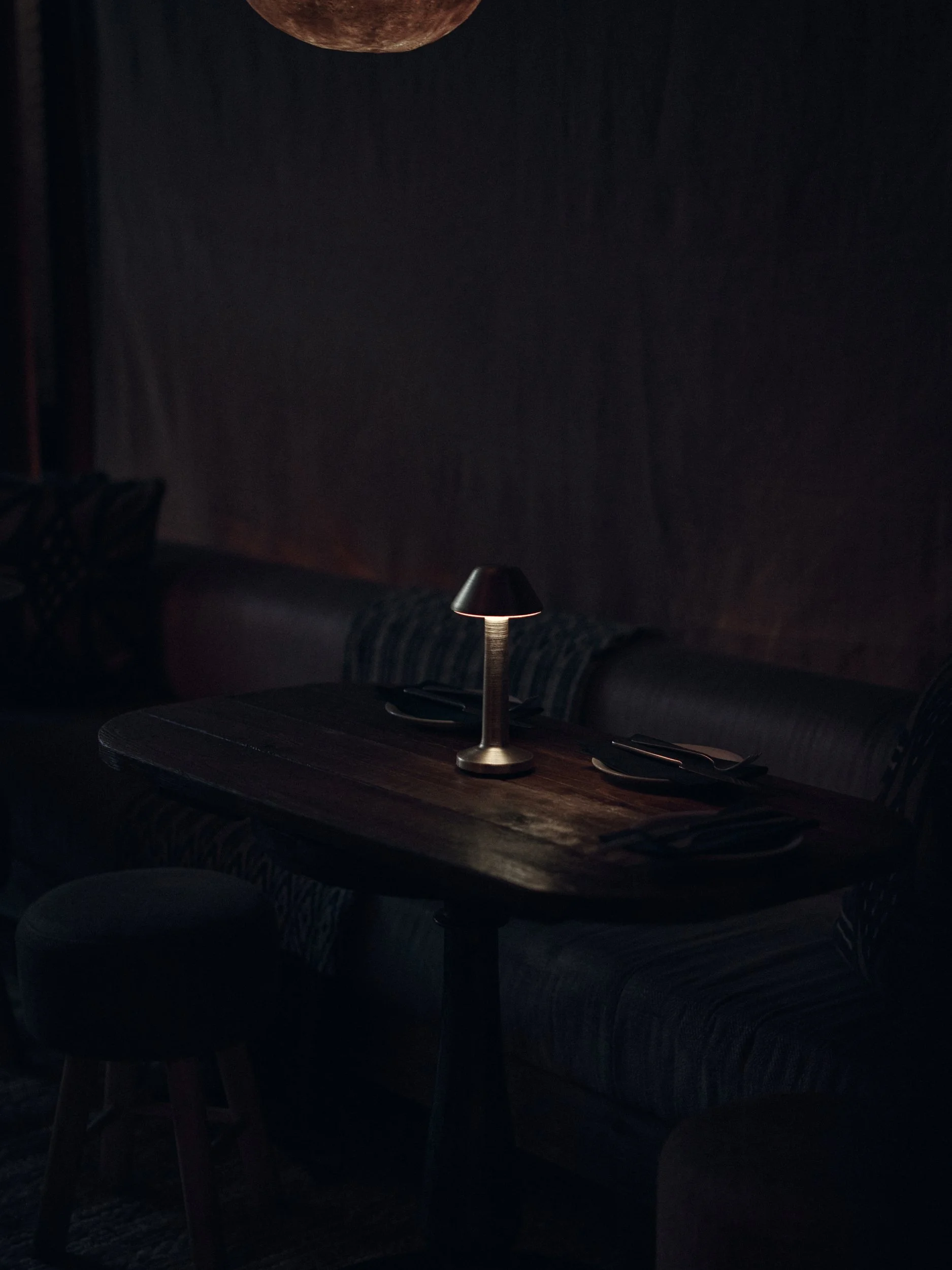 A dimly lit restaurant or lounge with a small round table. On the table, there are a small lamp, a plate, and some utensils. The background features a dark wall and a plush, patterned sofa.