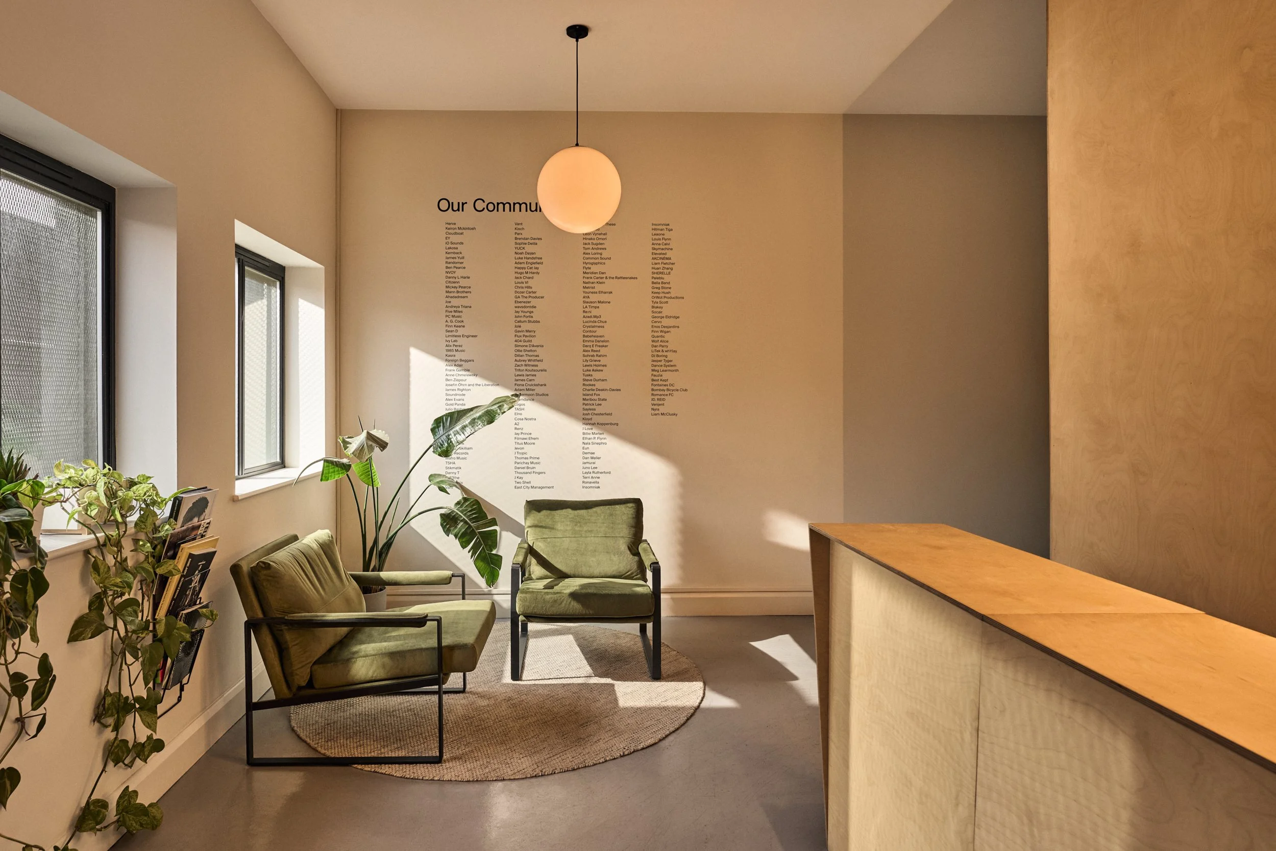 A cozy waiting area with two green armchairs, a large potted plant, and a wall with a list titled 'Our Community' featuring numerous names. There are windows on the left allowing natural light, and a beige and wooden reception desk on the right.
