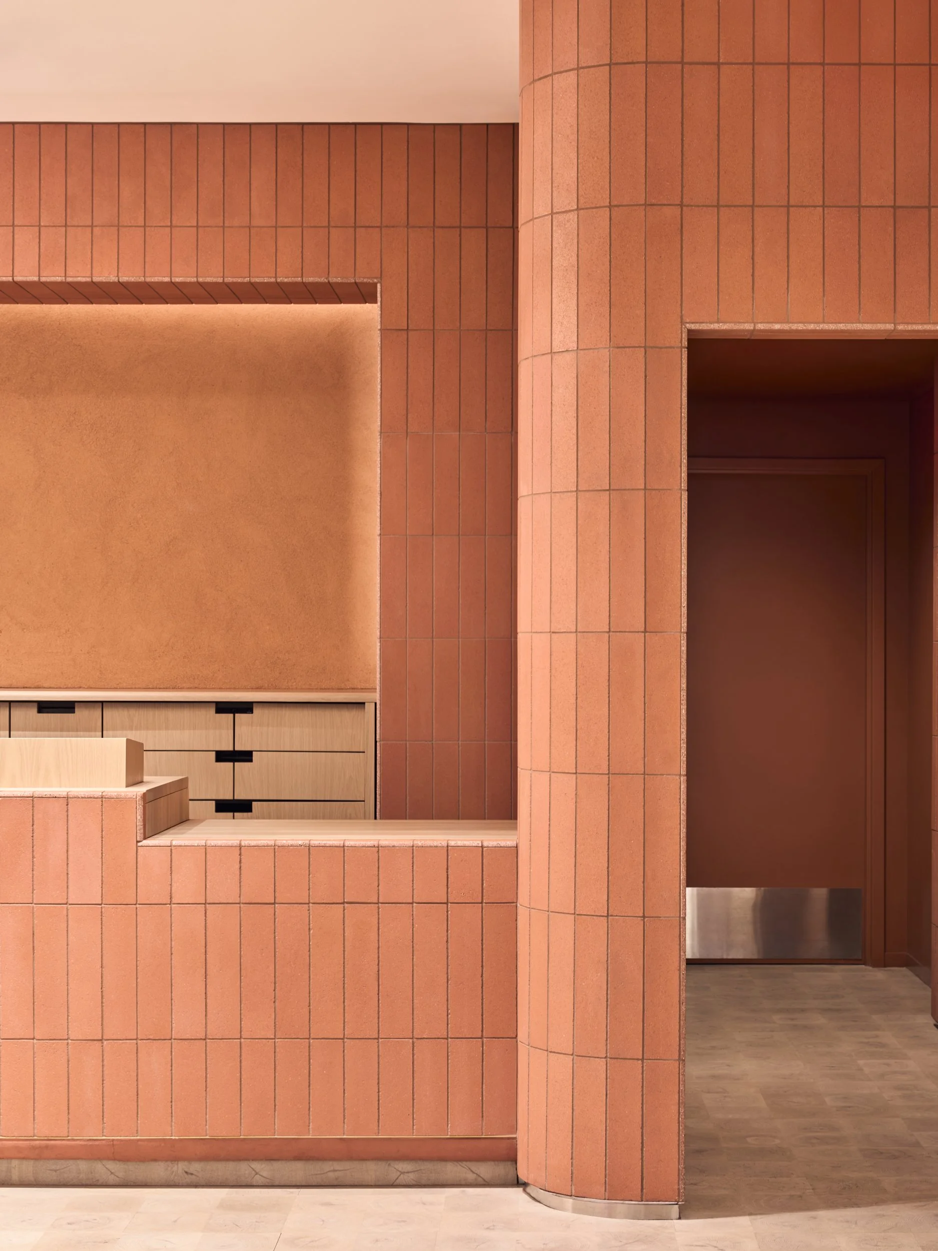 Interior view of a building with reddish-brown tiled walls, a wooden drawer, and a doorway leading to another room with wooden flooring.