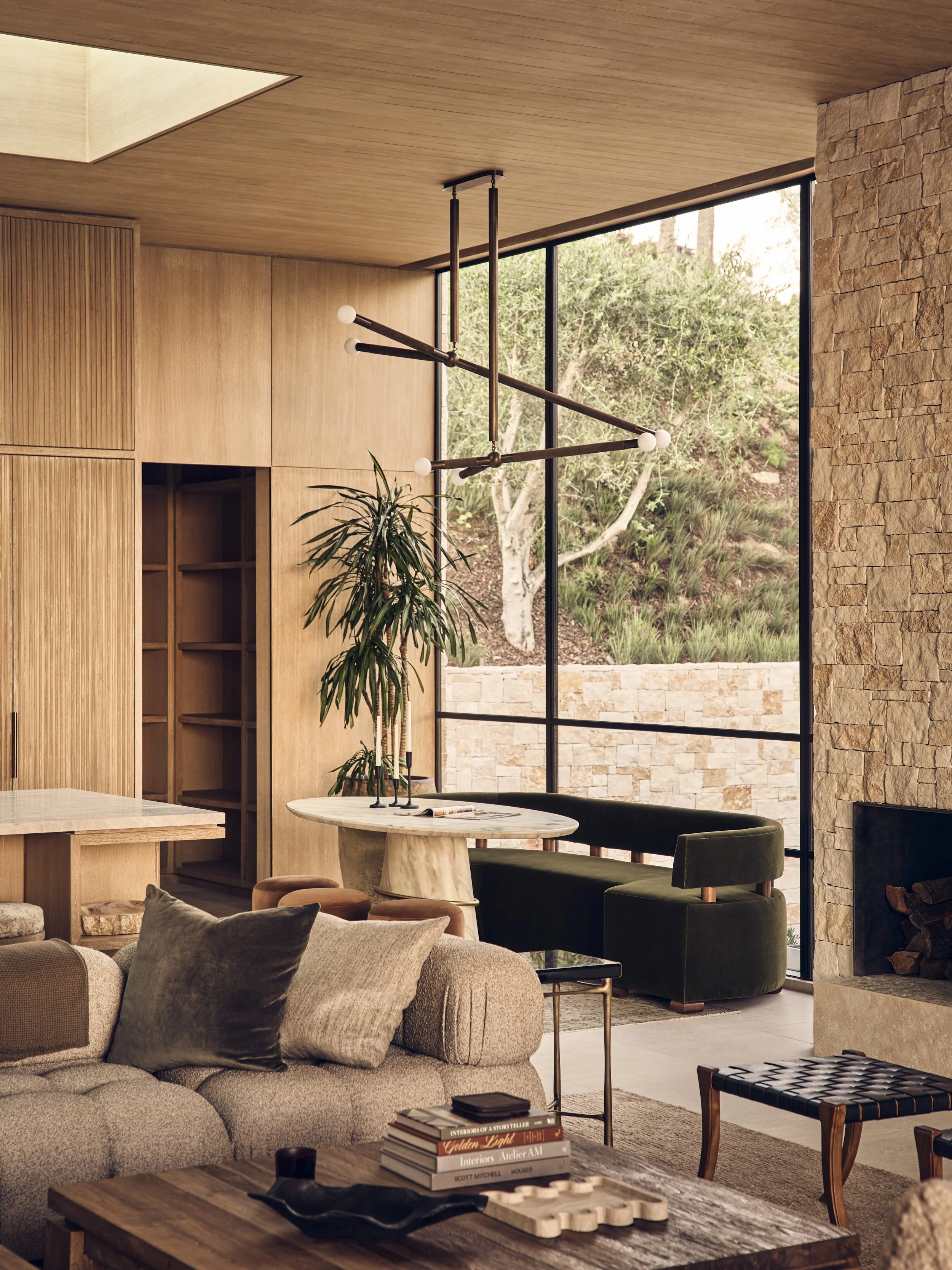 Modern living room with large floor-to-ceiling window featuring a view of trees and greenery outside. The space includes a beige curved sofa with dark throw pillows, a wooden coffee table with books and decorative items, a marble-top dining table wit
