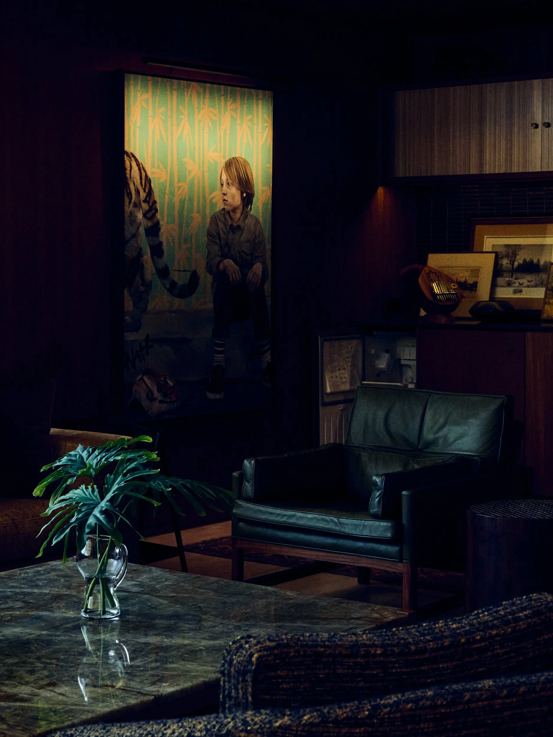 Living room with a green leather armchair, a glass table with a potted plant, and framed pictures on a wall. A painting of a woman and a cat is visible in the background.