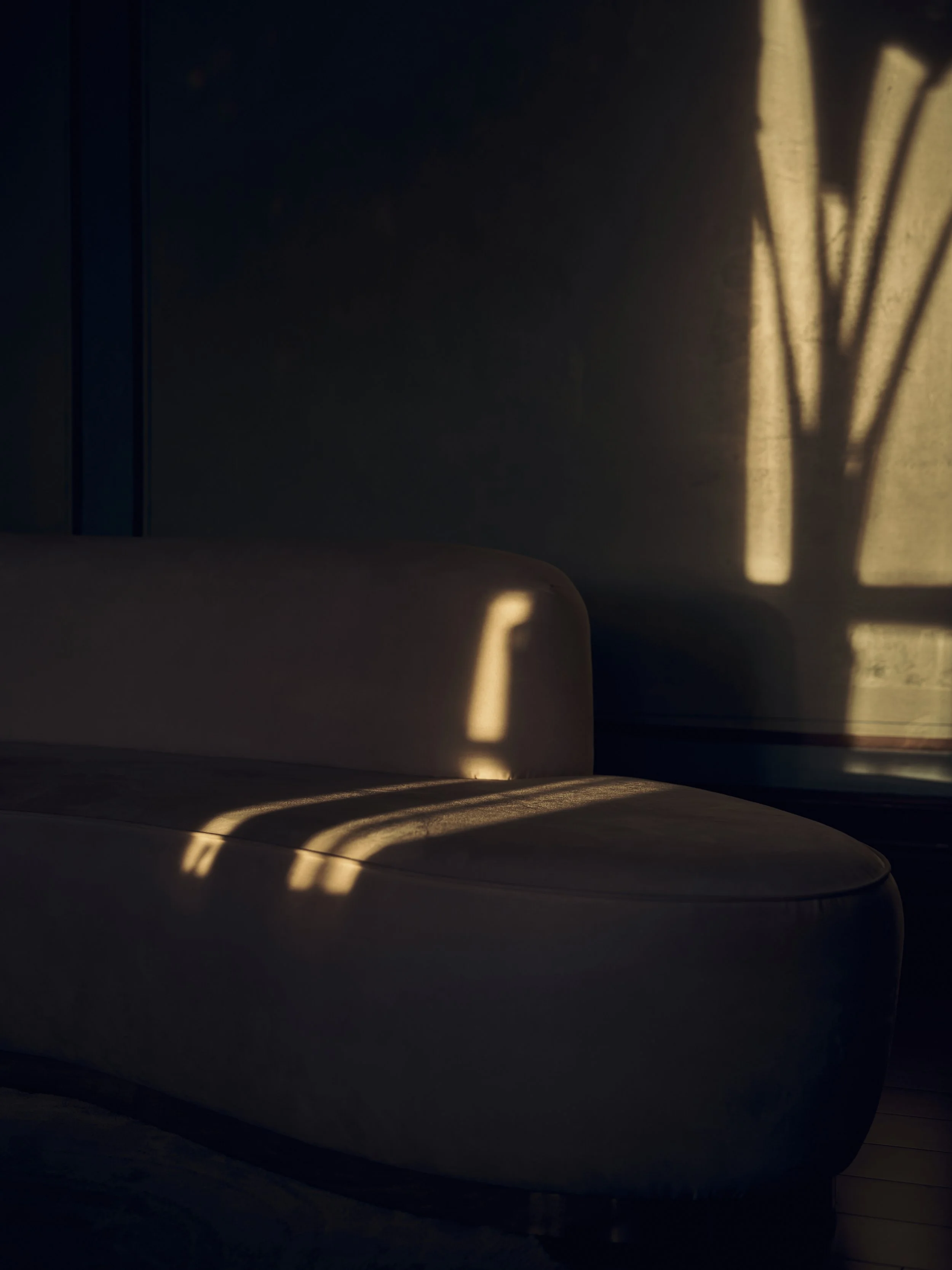 Sunlight casting shadows of window blinds onto a beige leather chair and a dark wall.
