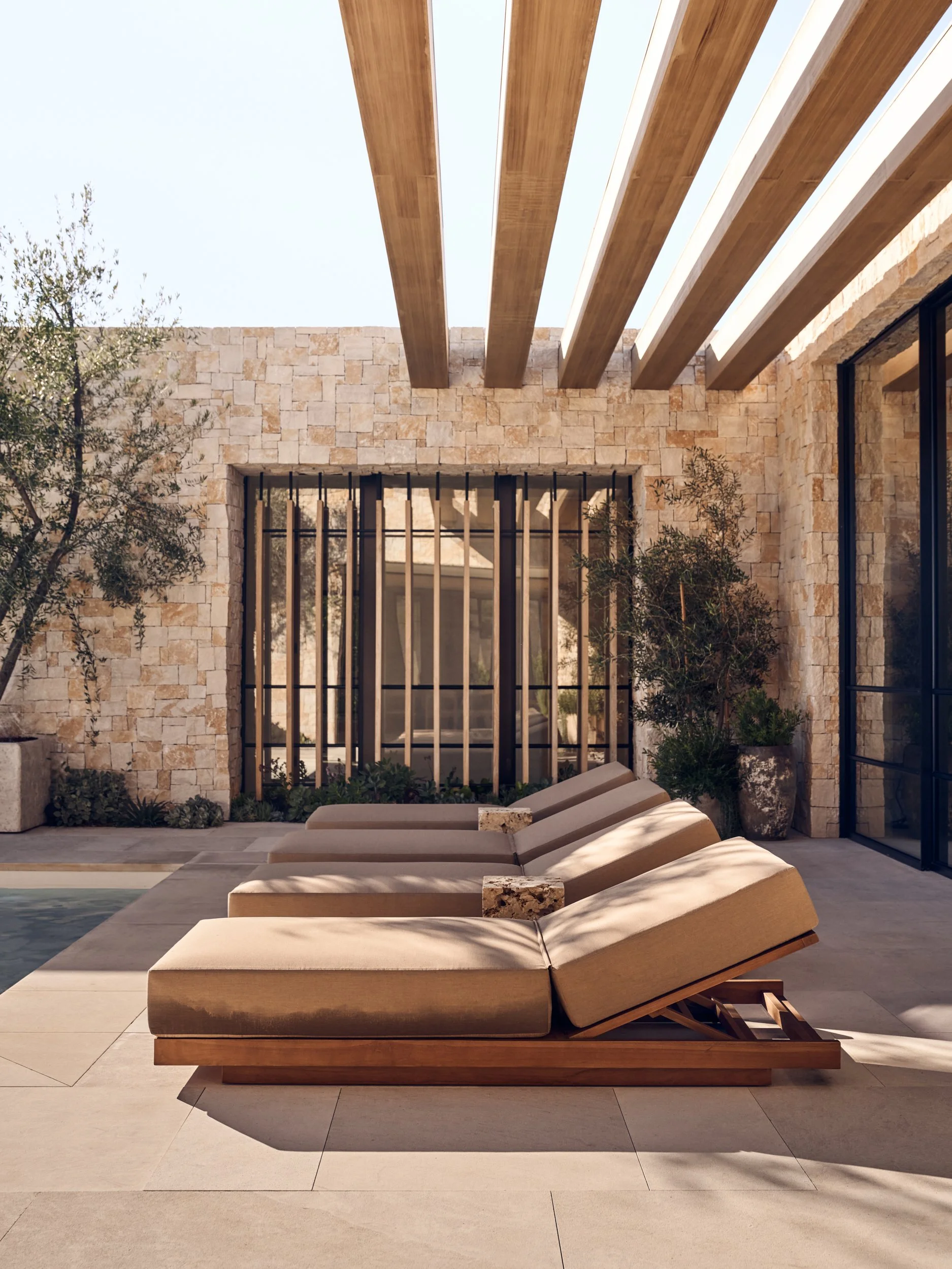 Outdoor patio area with beige cushioned lounge chairs, a stone wall with large rectangular windows, wooden beams overhead, and potted plants.
