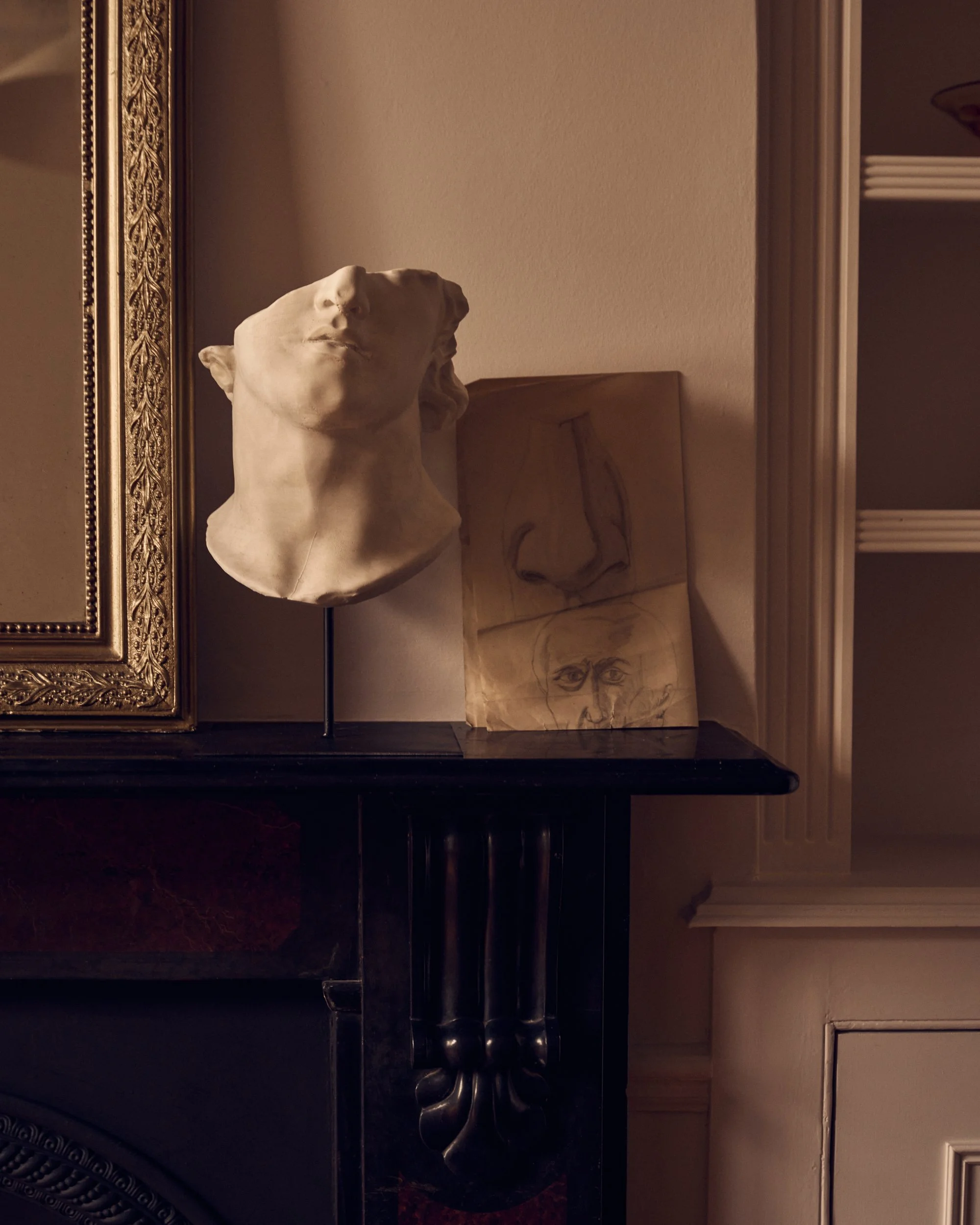 A white marble bust of a woman with curly hair, positioned on a black surface with a gold-framed mirror and sketches in the background.