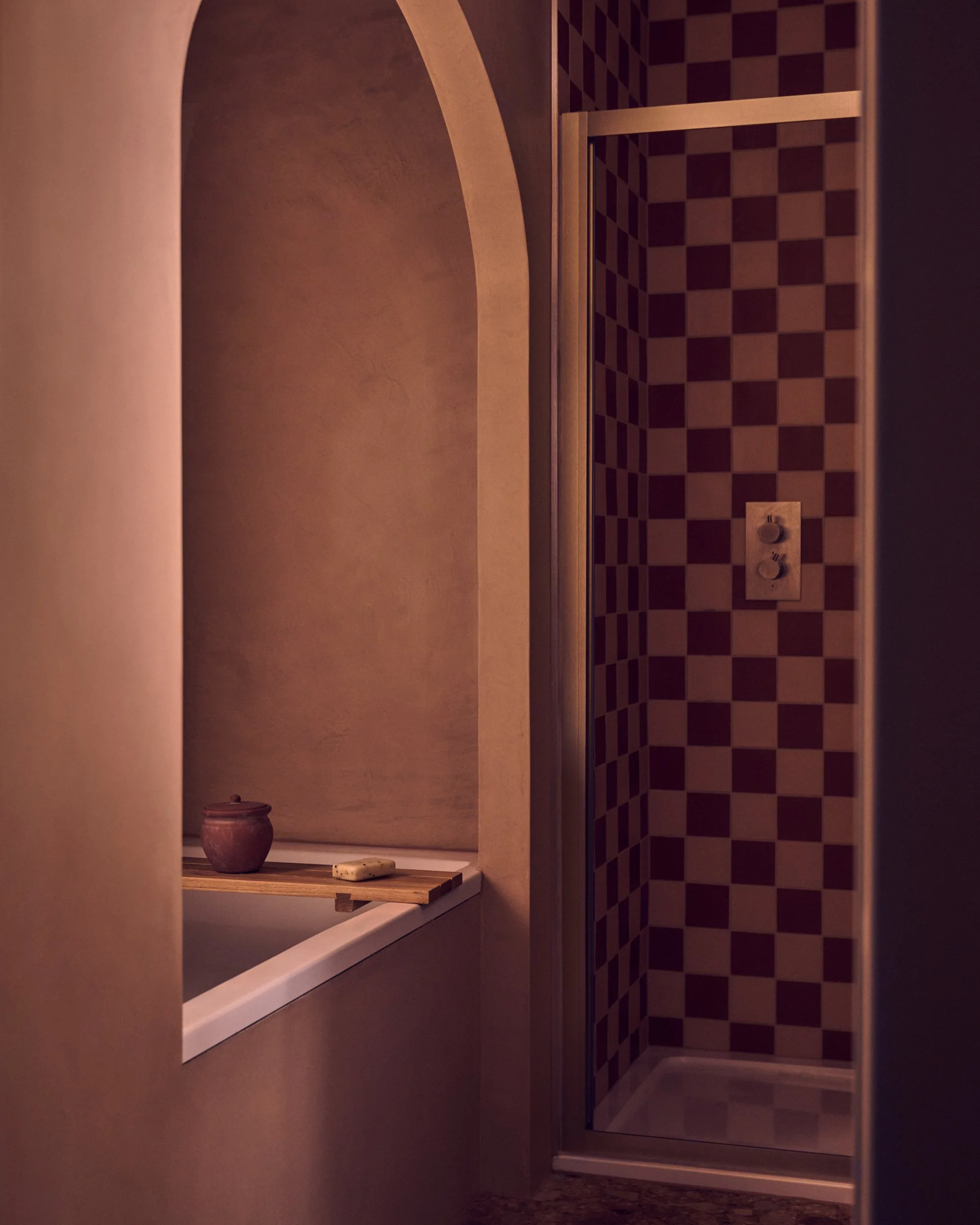 A bathroom shower with a checkered tile wall, a glass shower door with metal framing, a wall-mounted shower control, and a shelf with a small jar and soap, with a mirror reflecting part of the shower.