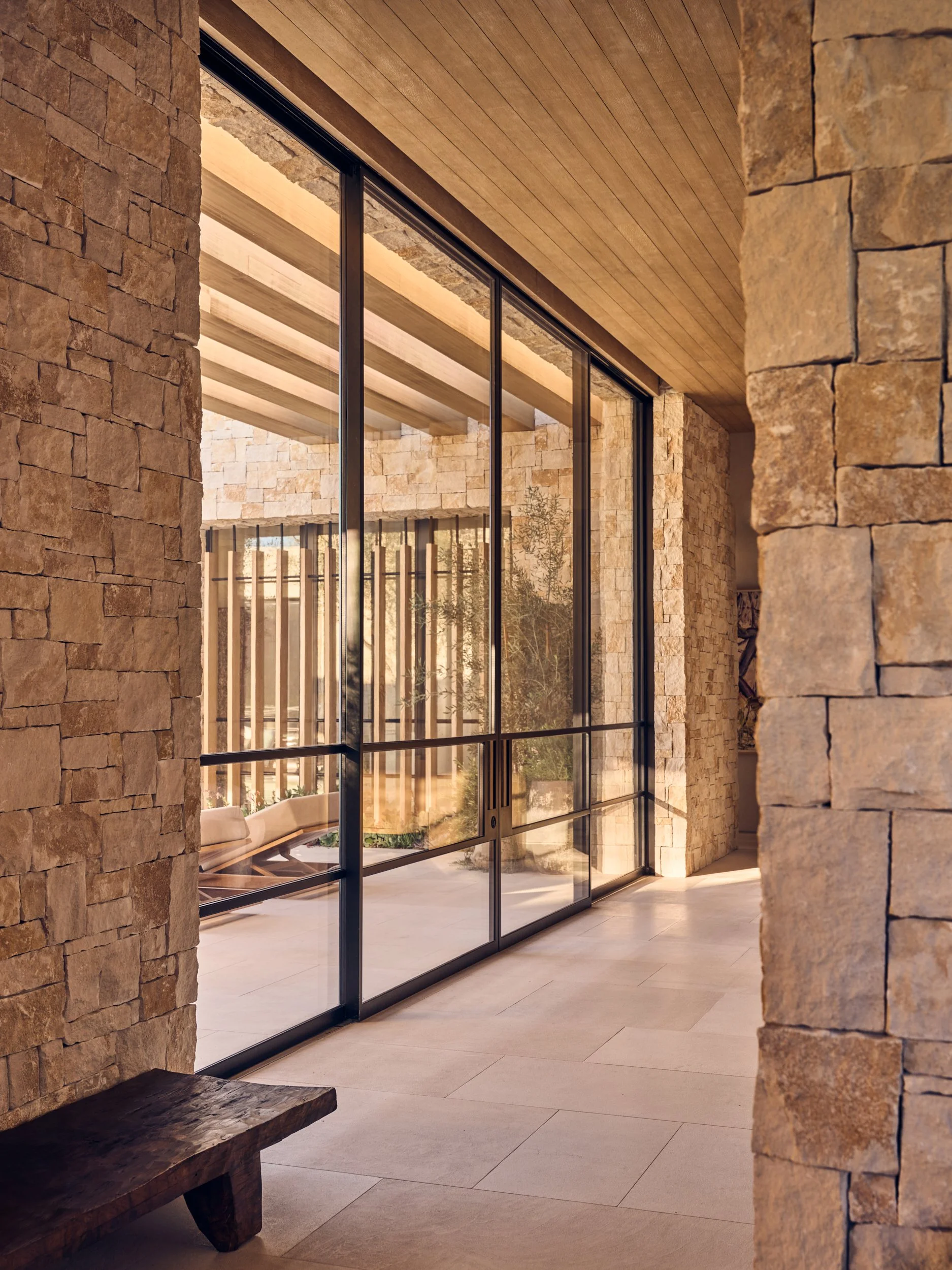Interior of a modern building with stone walls, wooden ceiling, large glass doors, and a bench.