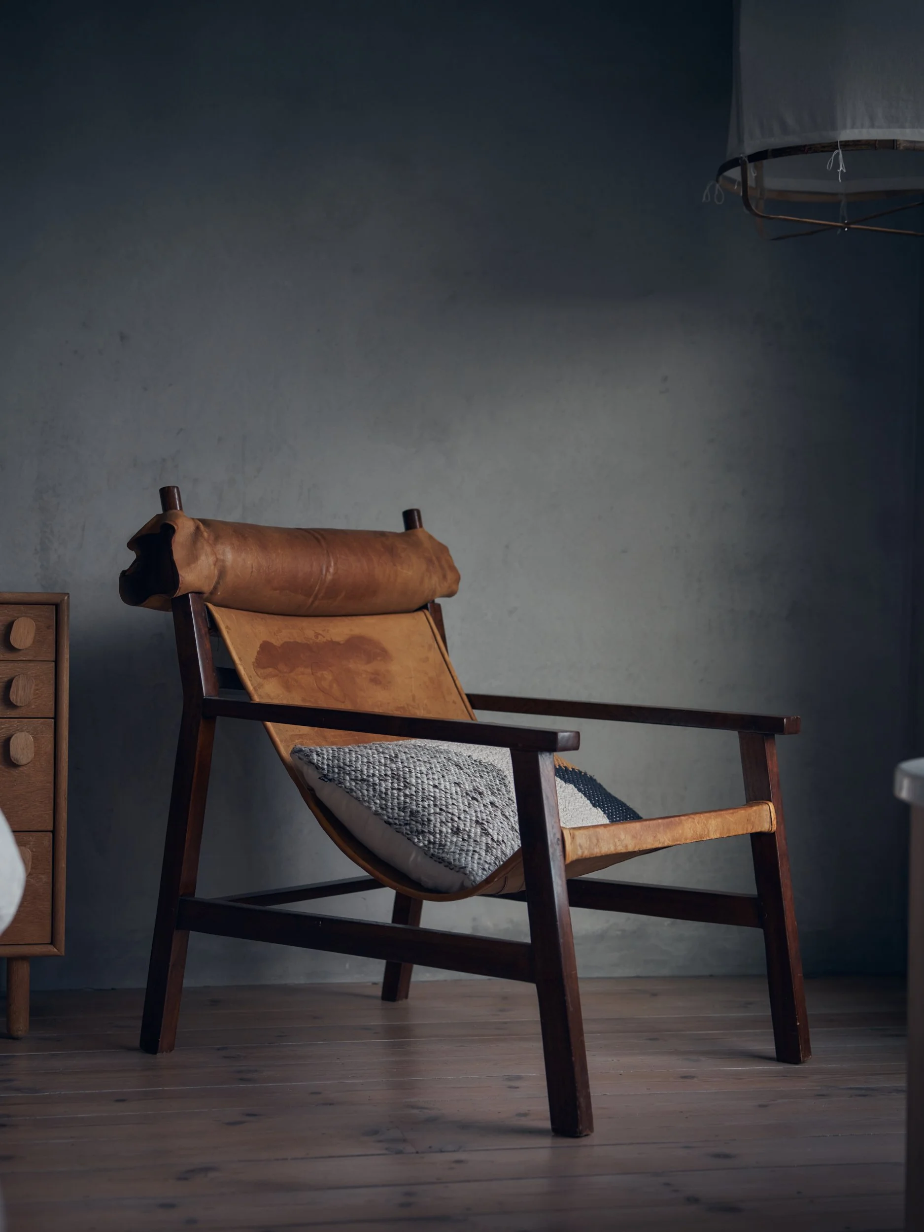 A wooden chair with a leather seat and a patterned cushion on the seat, positioned against a gray wall. Part of a small wooden cabinet is visible on the left, and a hanging paper lantern is partially visible on the top right.