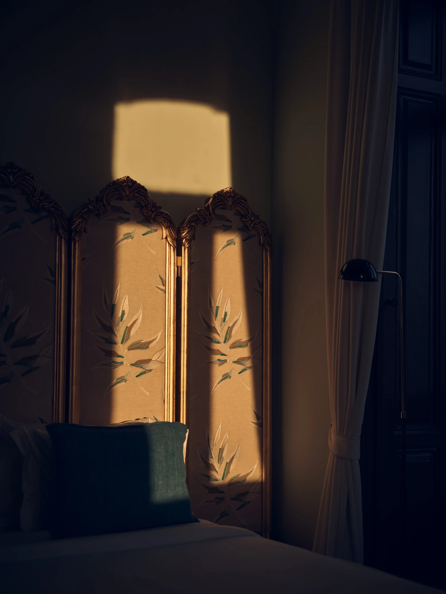 Sunlight casts a shadow of a window onto a beige, patterned room divider with embroidery, situated behind a bed with a gray pillow, in a dimly lit bedroom with a tall window and curtain on the right.