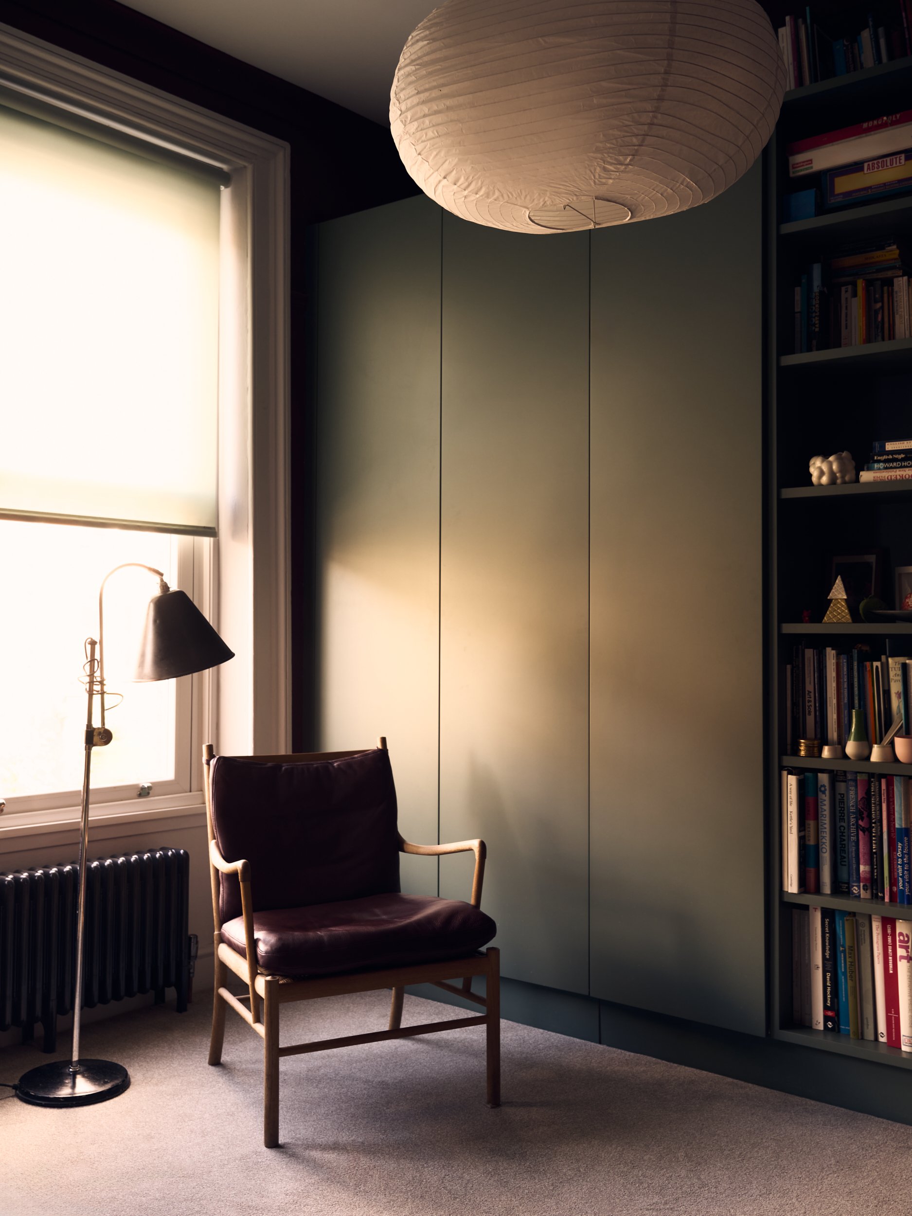 A cozy home office corner with a purple velvet armchair, a black floor lamp, a window with a white roller shade letting in natural light, a dark green closet or cabinet, and a tall bookshelf filled with books and decorative items, all on a beige carp