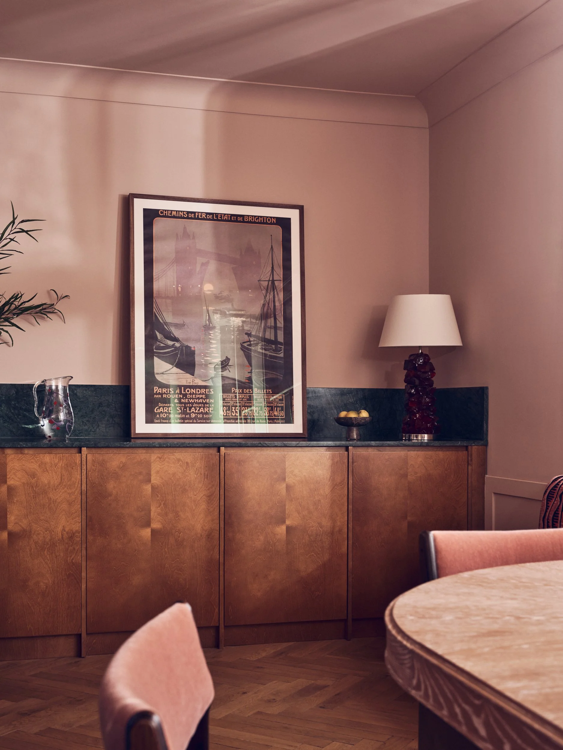 A room with wooden furniture, a framed poster on the wall, a table with a bowl of lemons, a lamp, and chairs.