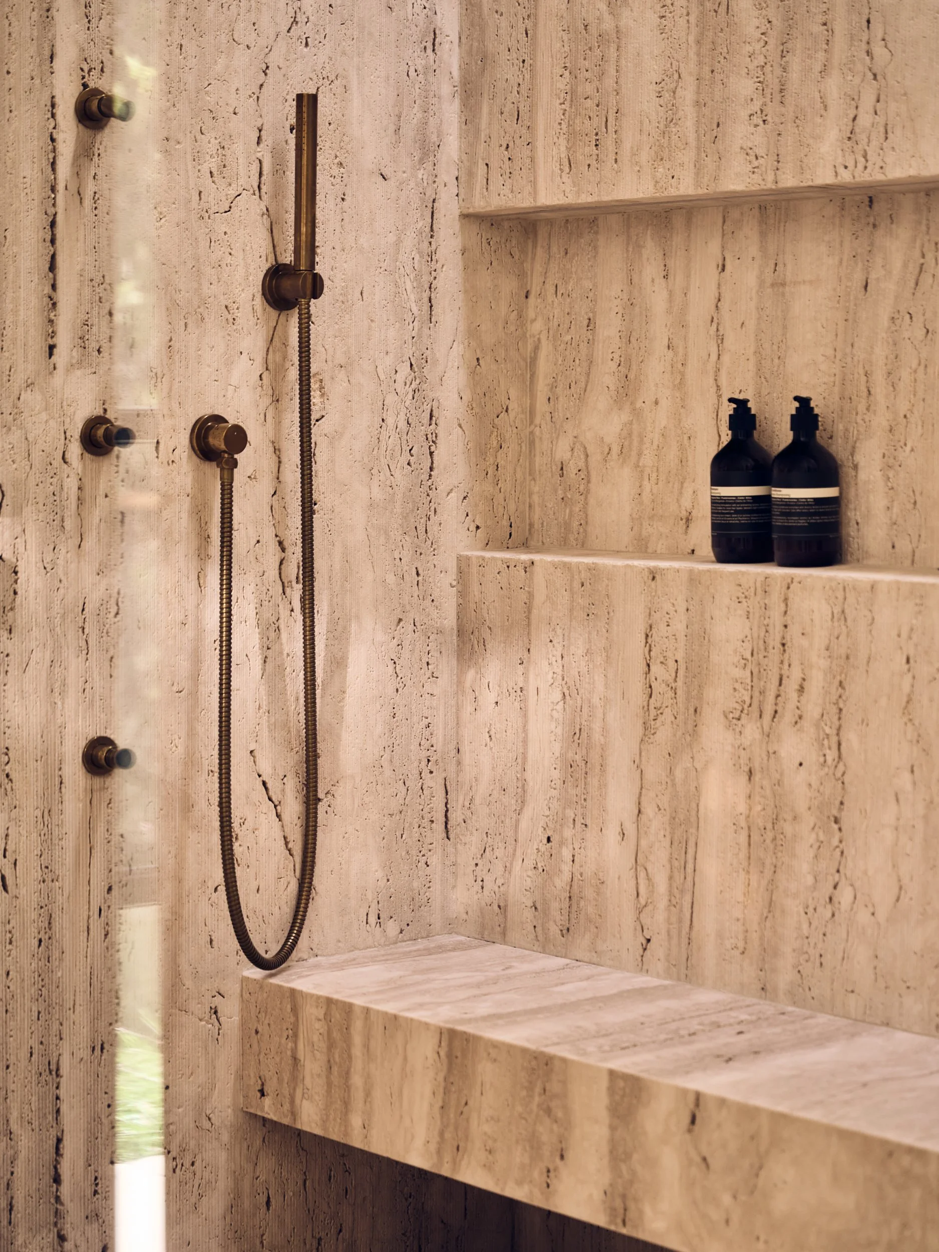 A bathroom shower area with beige stone tiles on the walls and built-in benches. There are two black bottles with pump dispensers on a ledge and a metal shower hose attached to the wall.