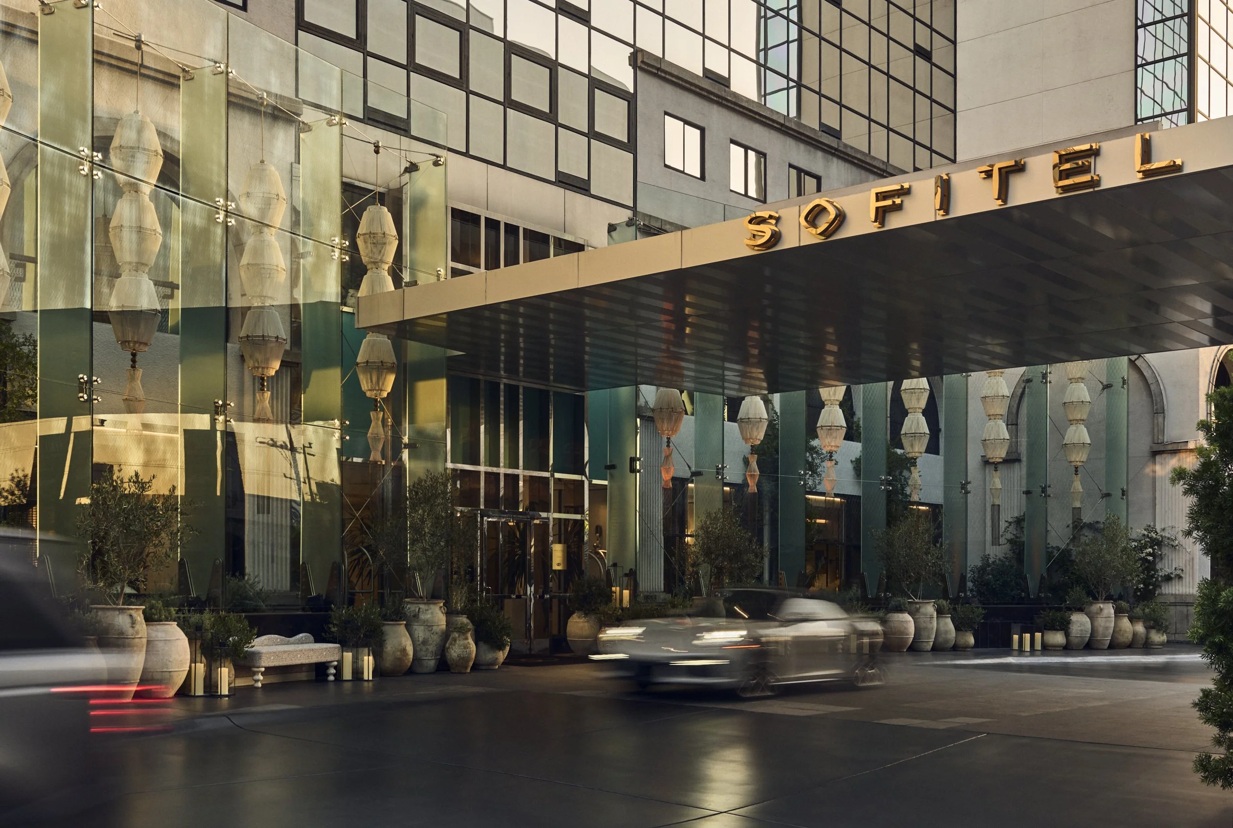 Modern building with glass and concrete architecture, illuminated signs, potted plants, and moving vehicles in front, reflecting a city street scene.