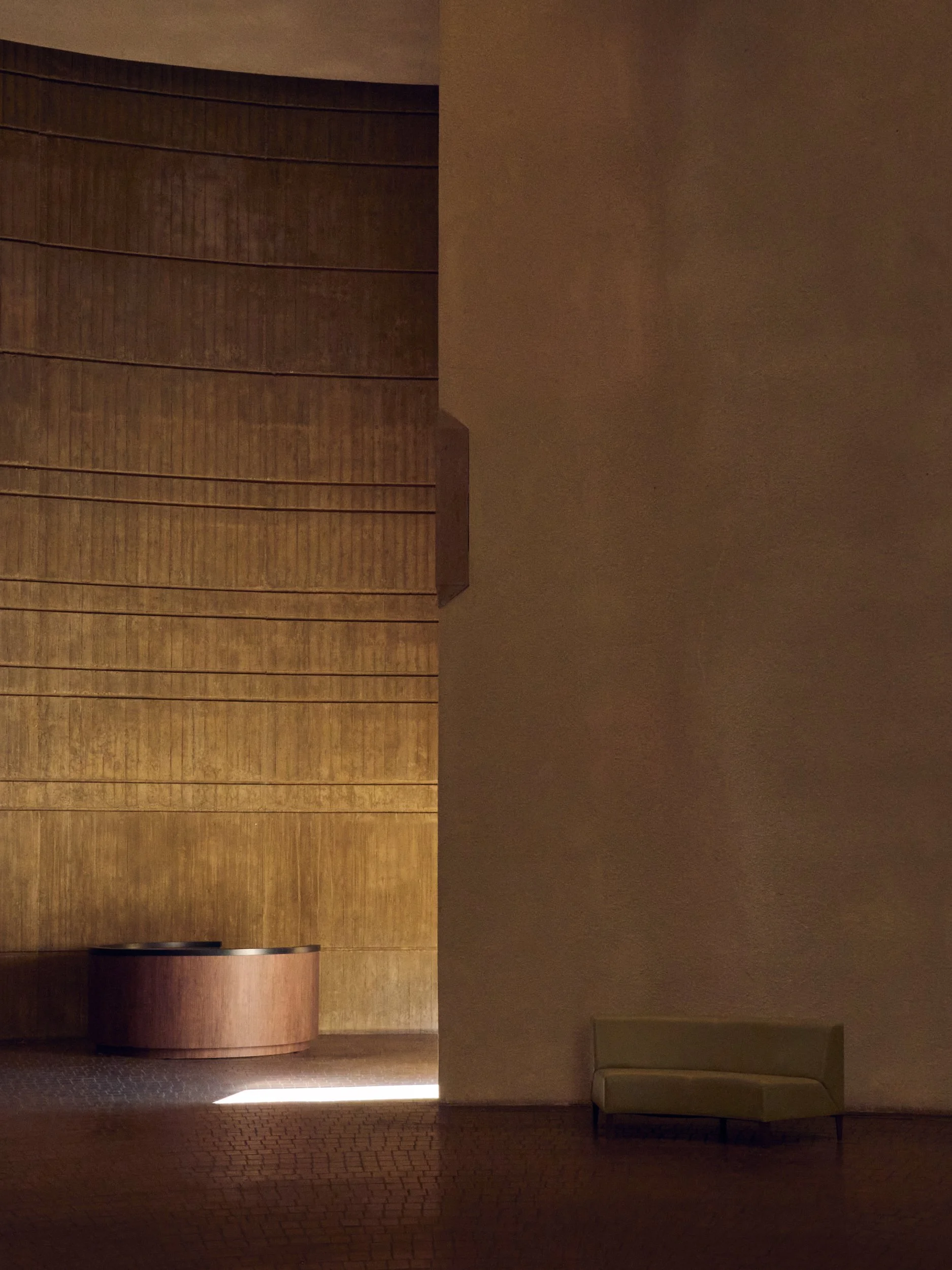 Dimly lit lobby with wooden wall panels, a curved wooden reception desk, and a beige sofa on the right side.