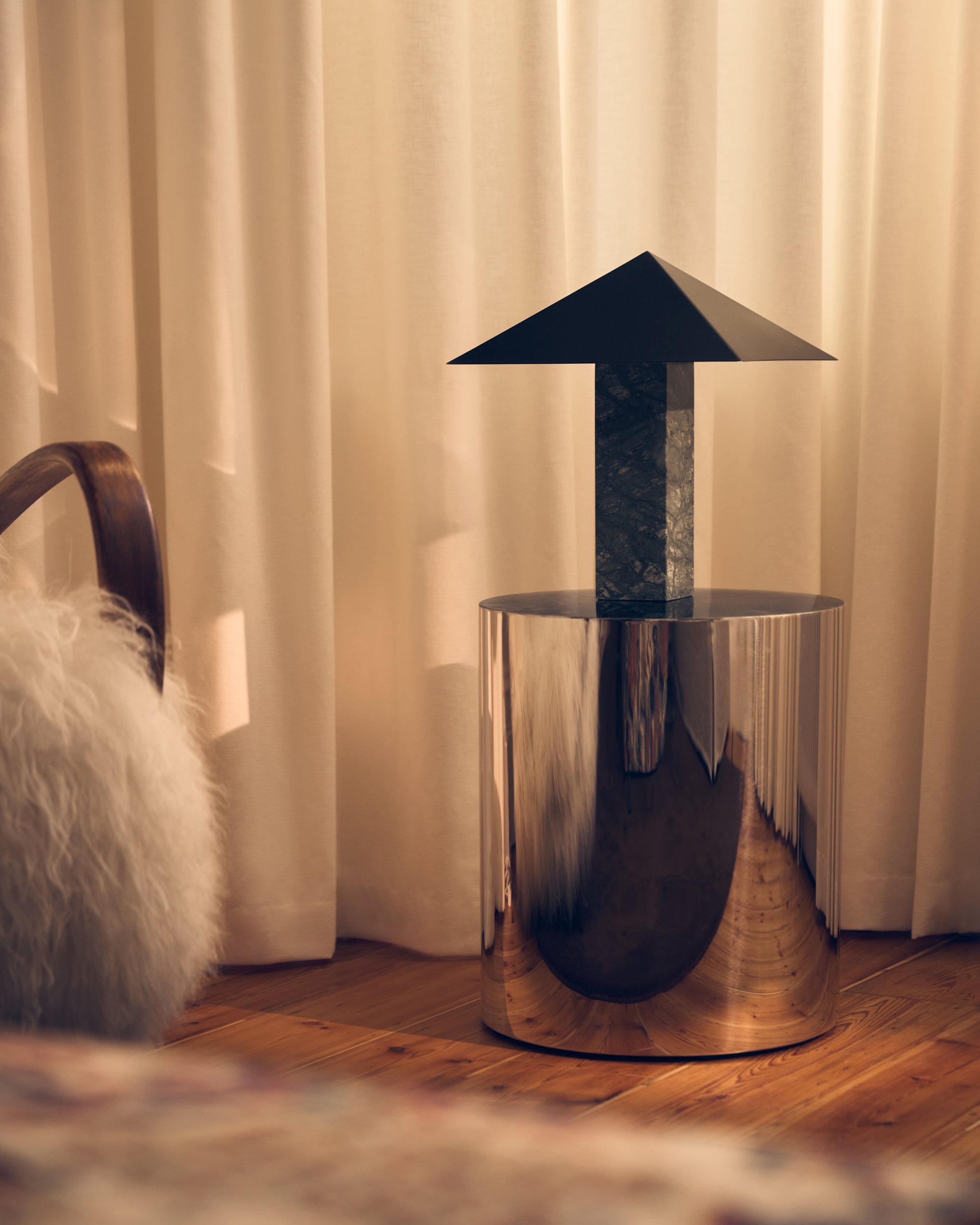 A dark-colored, geometric sculpture with a triangular top and rectangular base, placed on a shiny, reflective surface in front of beige curtains and a wooden floor.