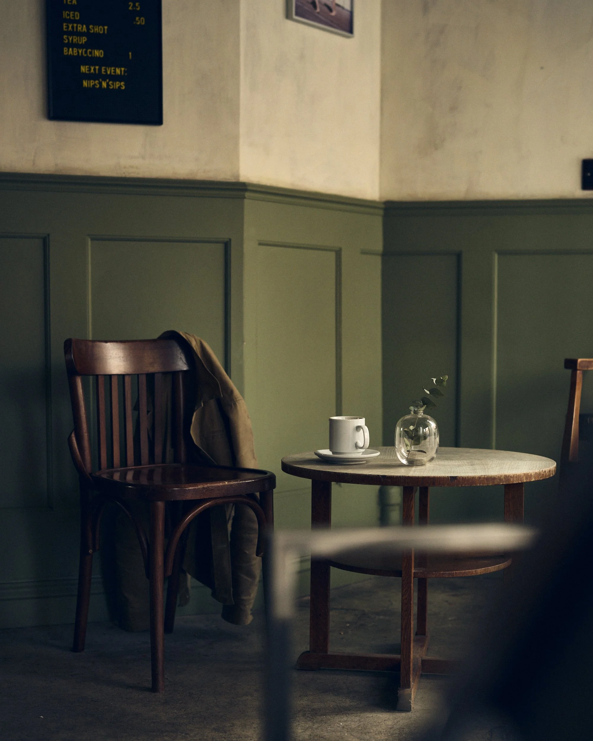 A cozy corner in a cafe with a round wooden table, a white mug, a small glass vase with eucalyptus leaves, and two wooden chairs, one with a jacket draped over it. The background features green wall paneling and a coffee menu sign.