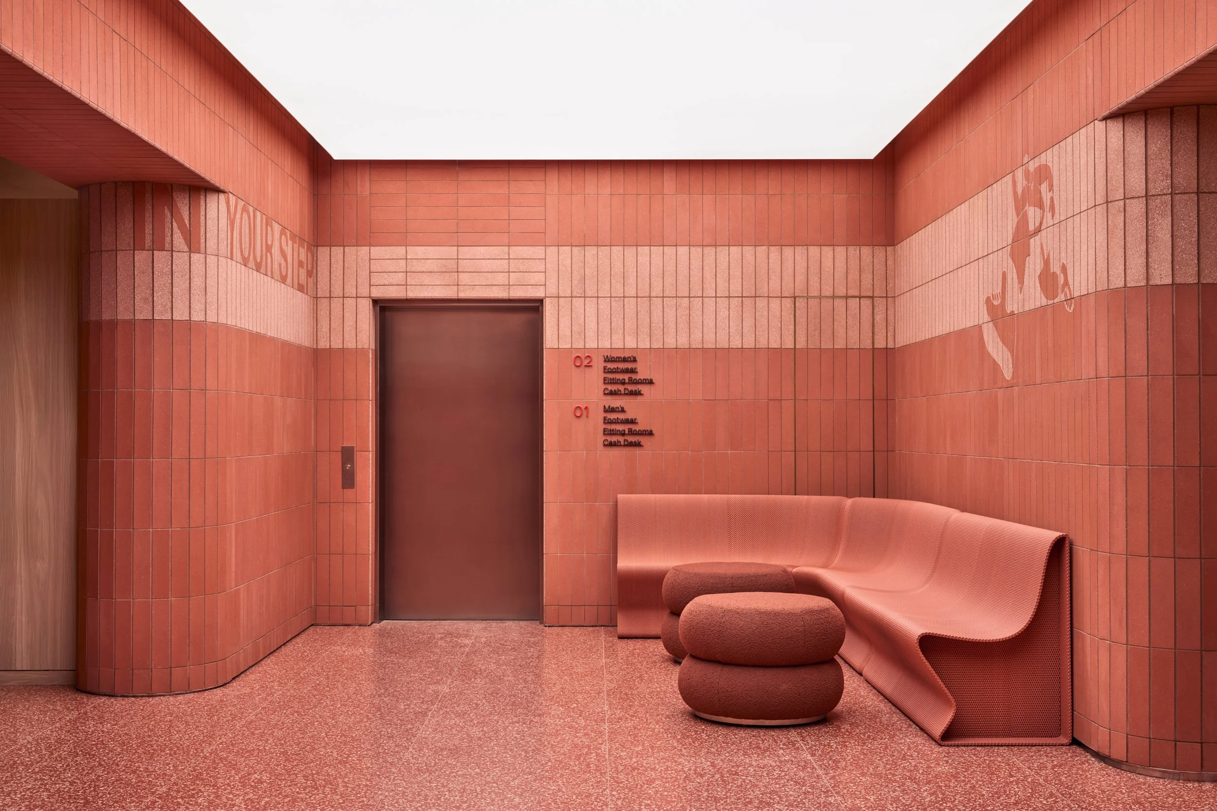 A modern wall with pink tiles and pink seating with curved design, elevator door in the center, signage for women's and men's restrooms, and stylized pink and white text on the wall.