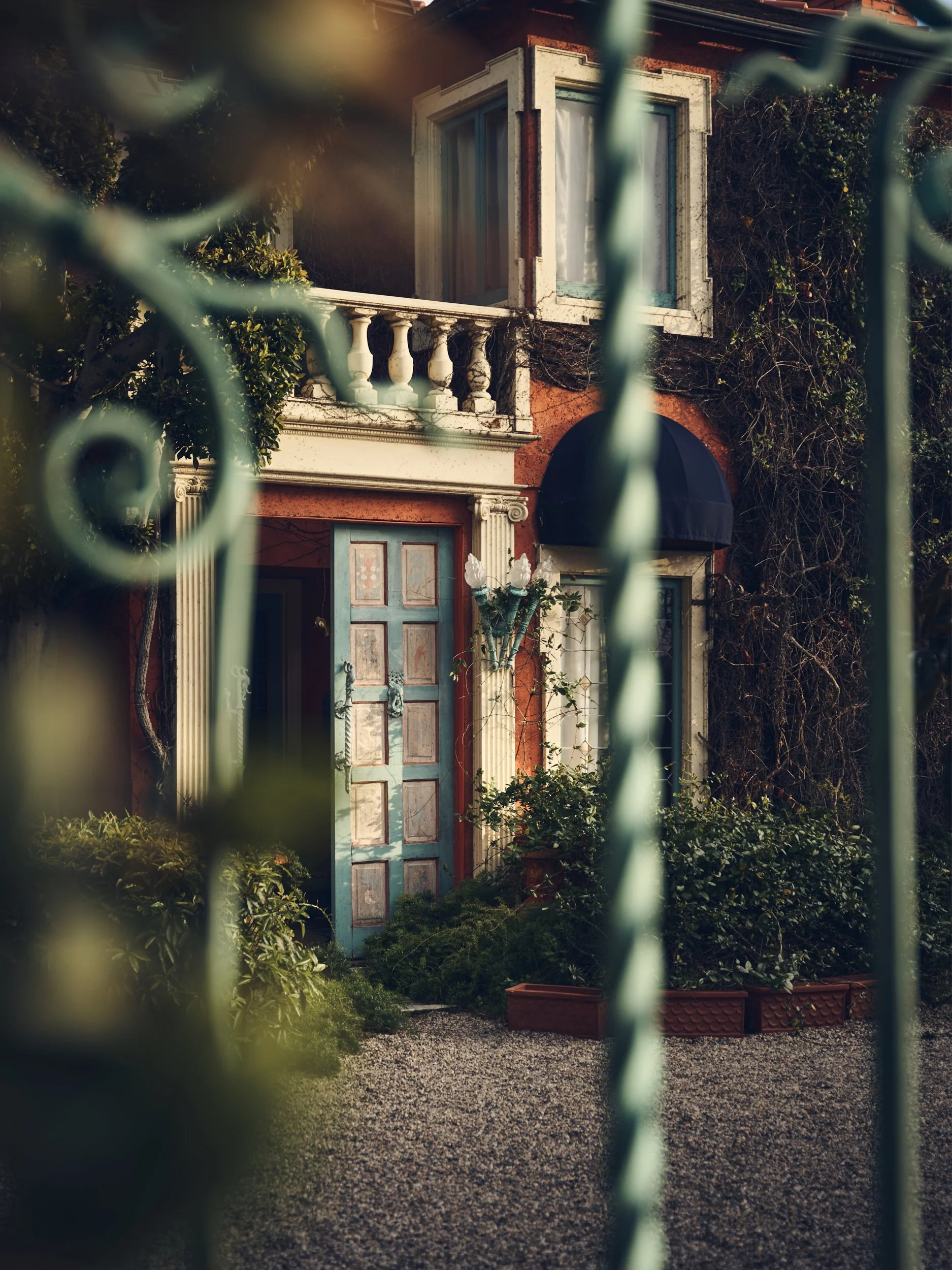 View of a house's front entrance through a decorative wrought iron gate, showing a blue door, an upper-floor window, a black awning, and garden greenery.