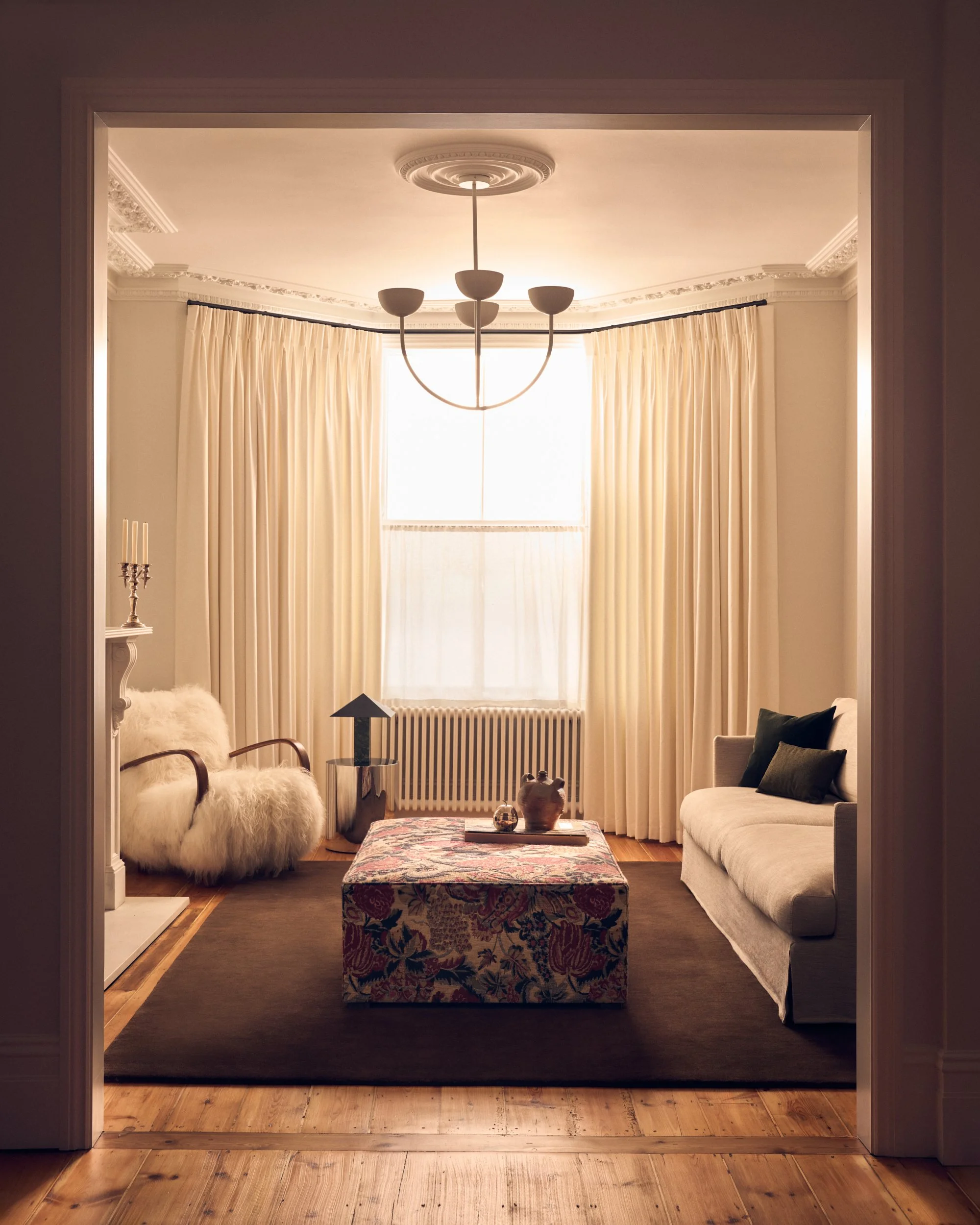 Living room with cream curtains, a white sofa with dark pillows, a floral ottoman, a side table with decorative items, and a chandelier in a bright space with wooden floors.