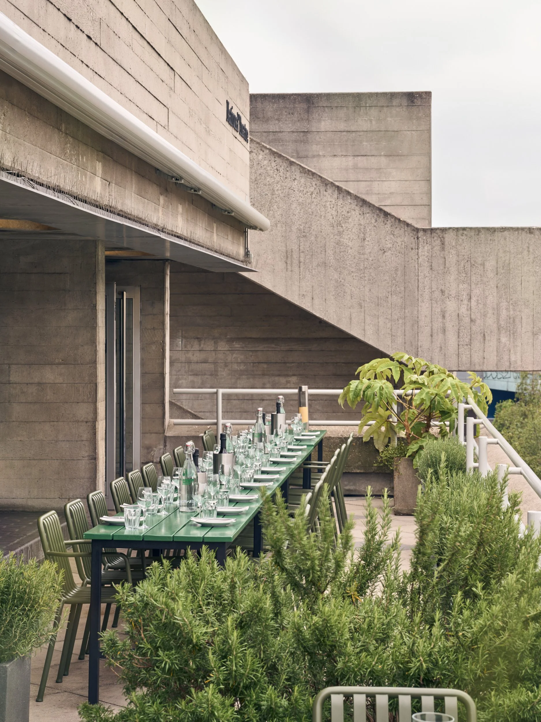 Outdoor dining area with a long green table set with glasses and bottles, surrounded by green plants, against a concrete building with modern architectural style.